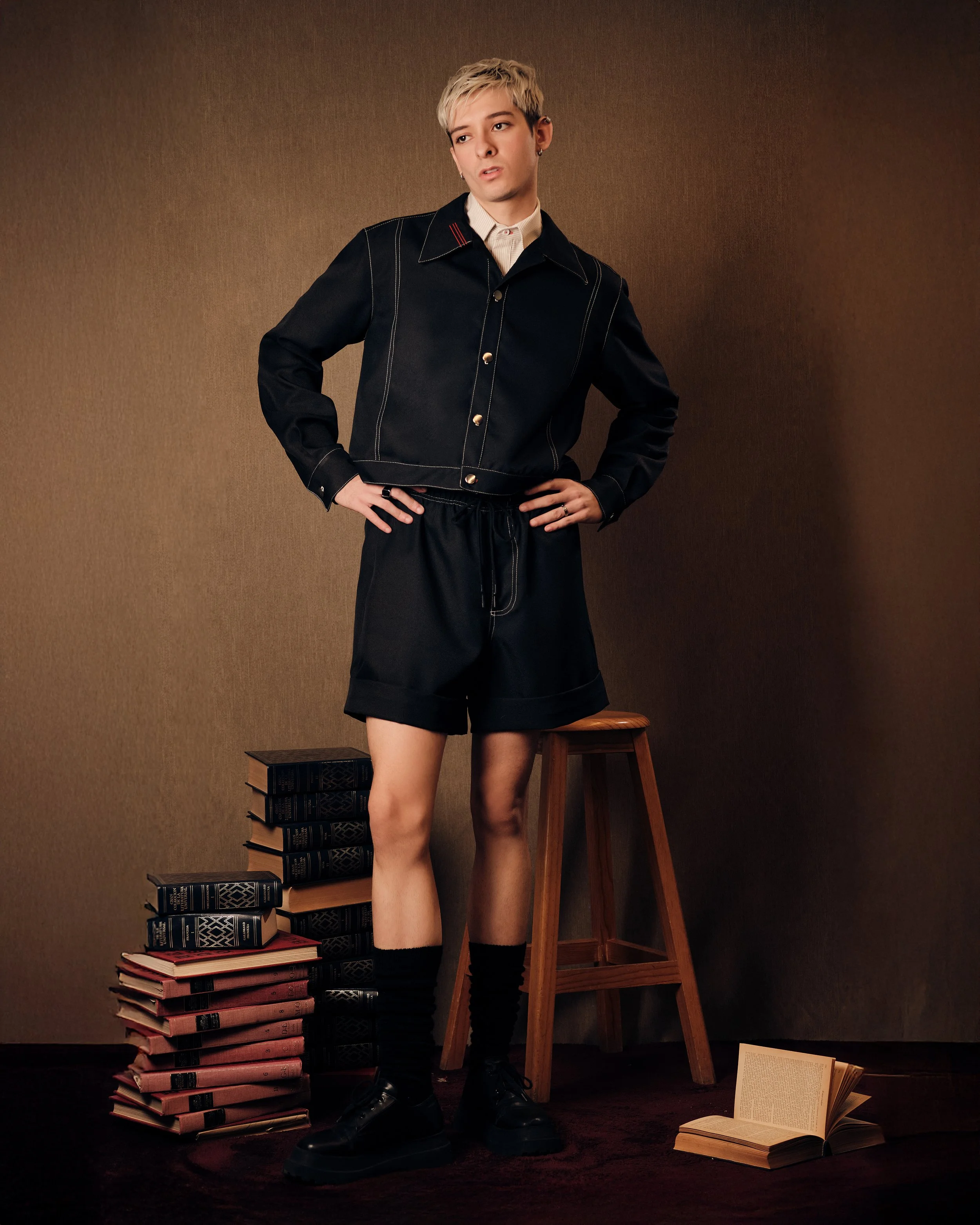 Joven con ropa de estilo casual en un fondo marrón, rodeado de pilas de libros apilados en el suelo y un libro abierto en el piso.