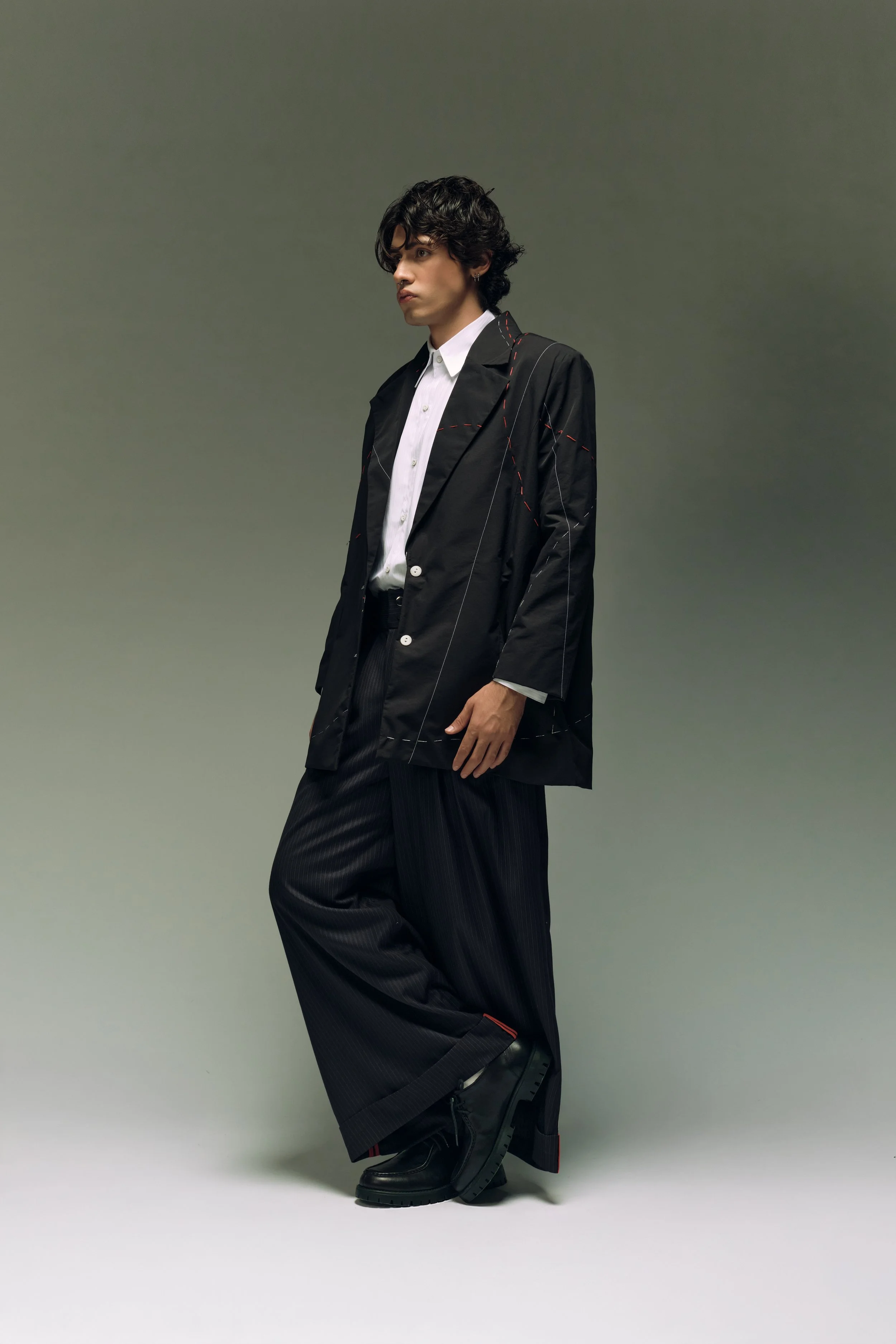 Hombre joven con cabello rizado oscuro, equipado con chaqueta negra, camisa blanca, pantalones anchos negros y botas negras con suela gruesa, posando sobre fondo gris liso.