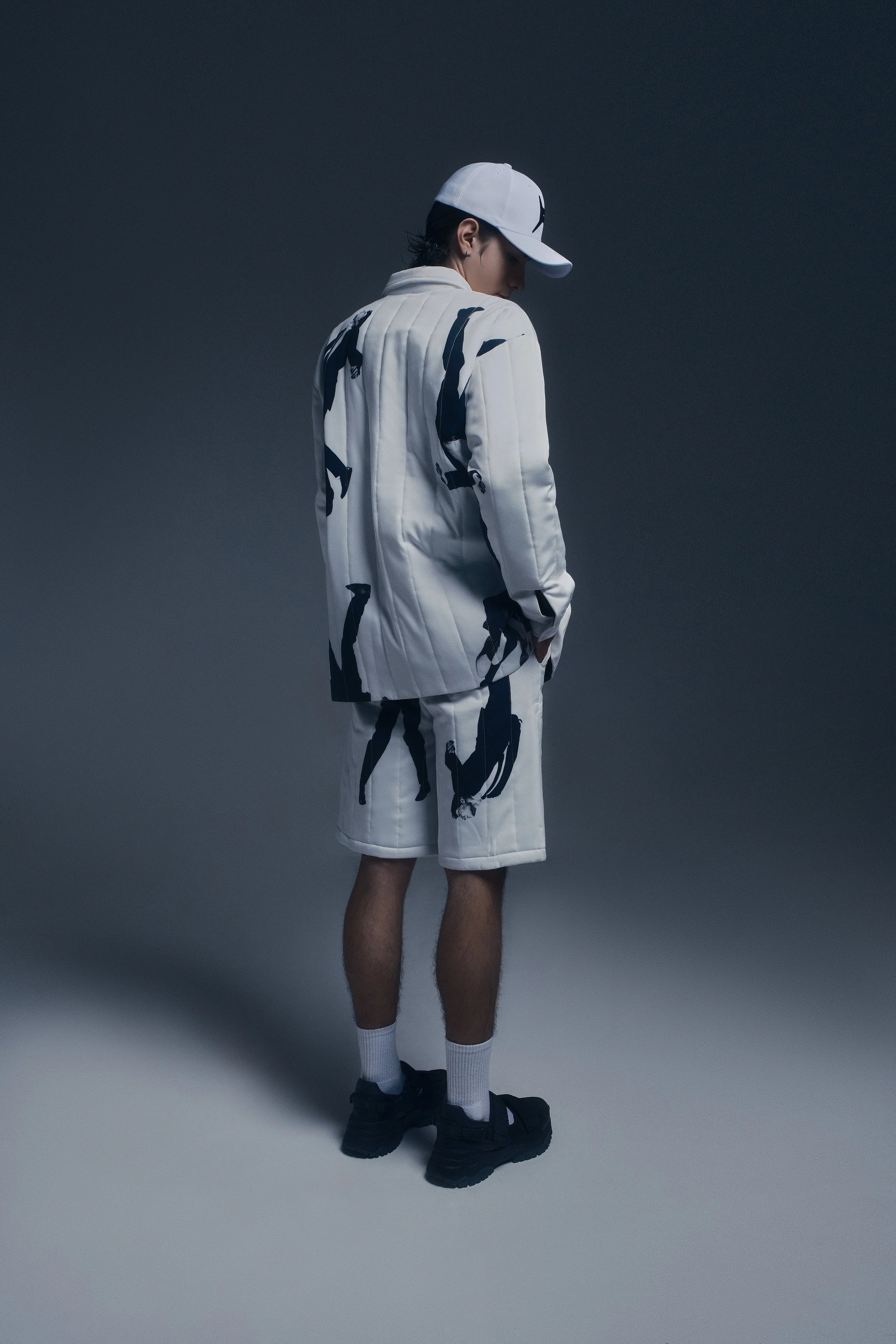 Persona usando ropa deportiva blanca con diseños en negro, gorra blanca y zapatos deportivos negros, de espaldas, en un fondo oscuro.