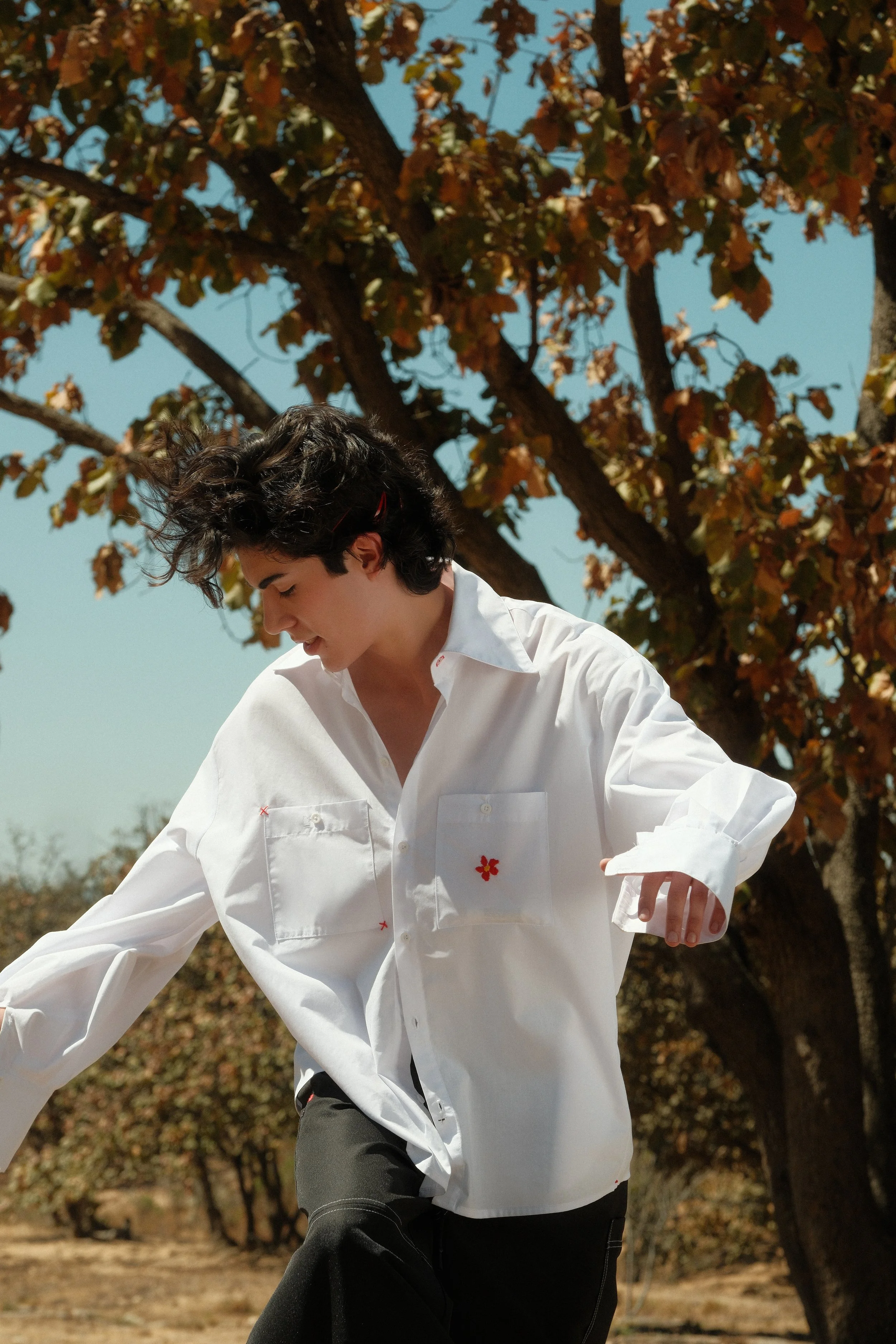 Joven con cabello oscuro y suelto, viste una camisa blanca con detalles rojos y está al aire libre junto a un árbol con follaje en tonos otoñales.