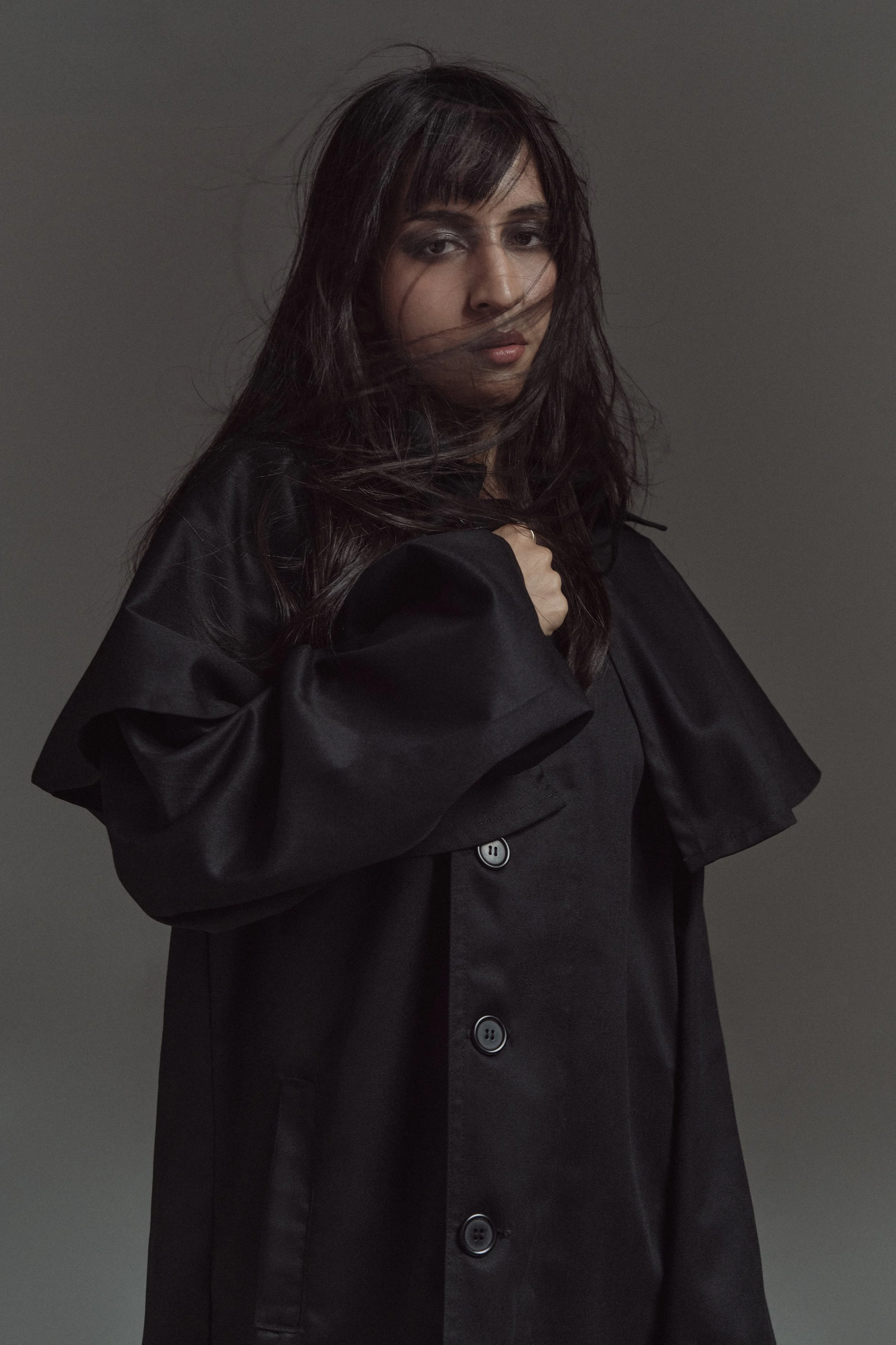 Mujer con cabello largo y oscuro, vestida con abrigo negro y blusa negra, en un fondo gris oscuro.