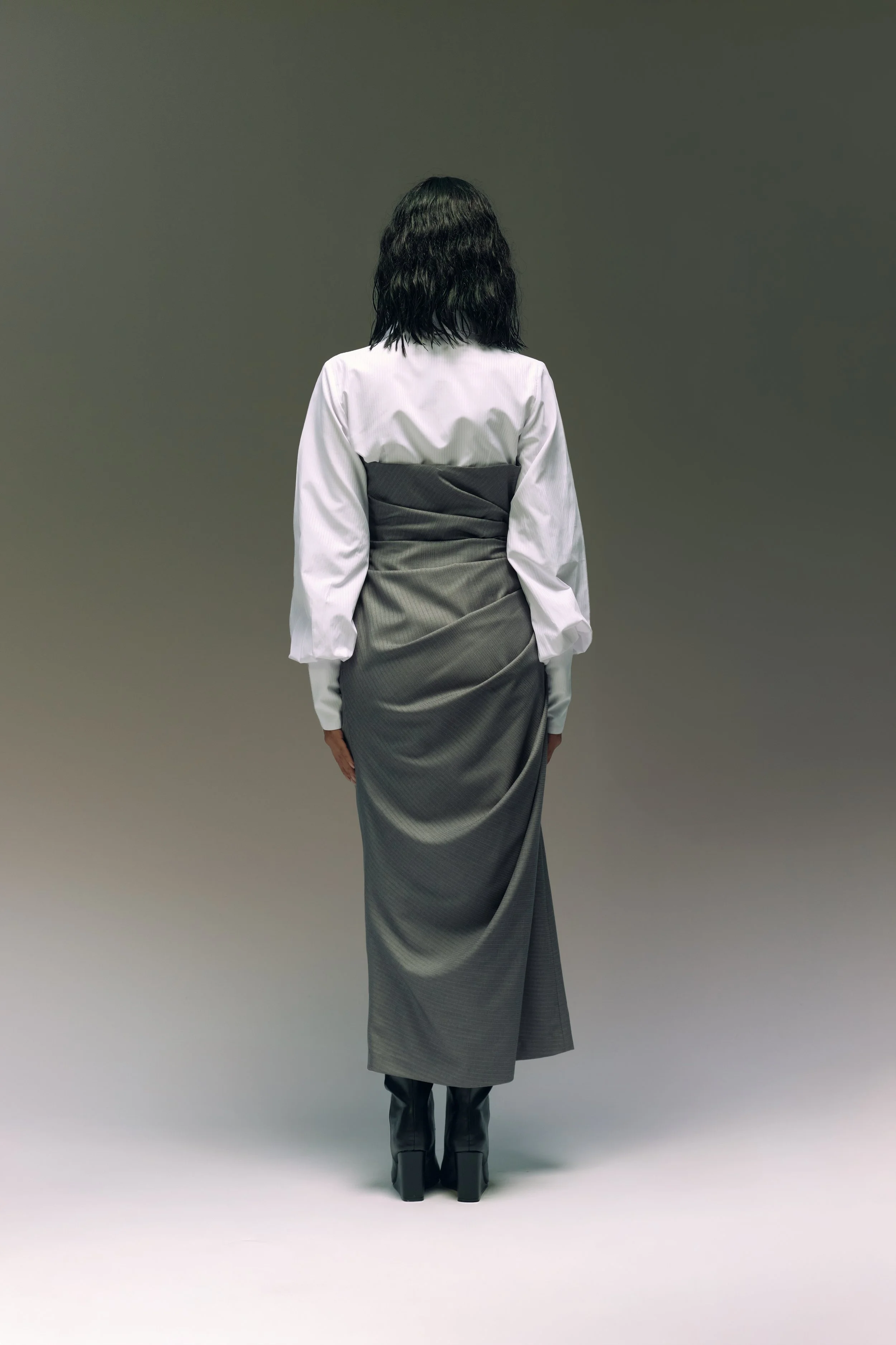 Persona de espaldas, con cabello oscuro, usando blusa blanca, falda larga gris y botas negras, en un fondo gris