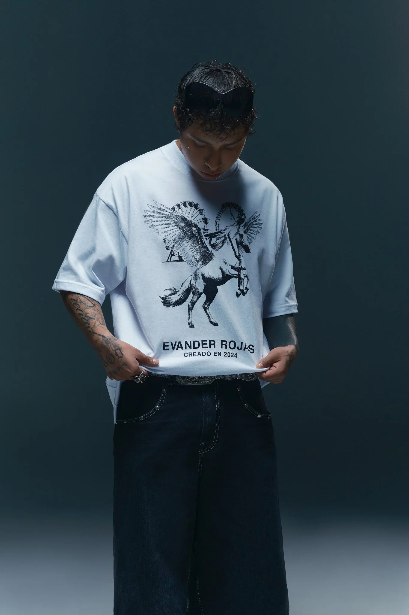 Joven con camiseta blanca con dibujo de un unicornio y palabras en español, pantalones oscuros, tatuajes en ambos brazos, gafas negras en la cabeza, fondo oscuro.