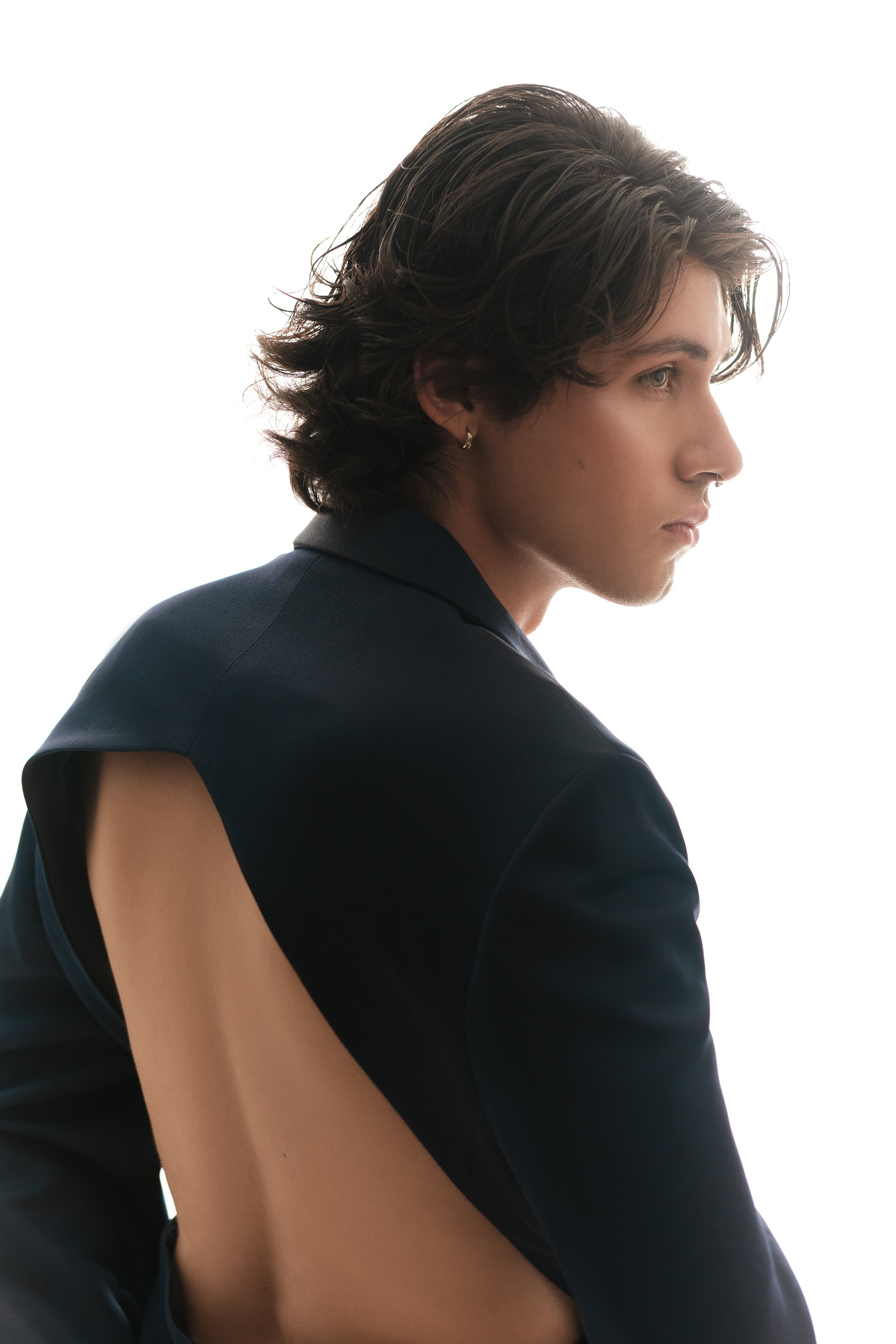 Retrato de un joven con cabello castaño oscuro y peinado de lado, usando abrigo negro con diseño abierto en la espalda, en fondo blanco.