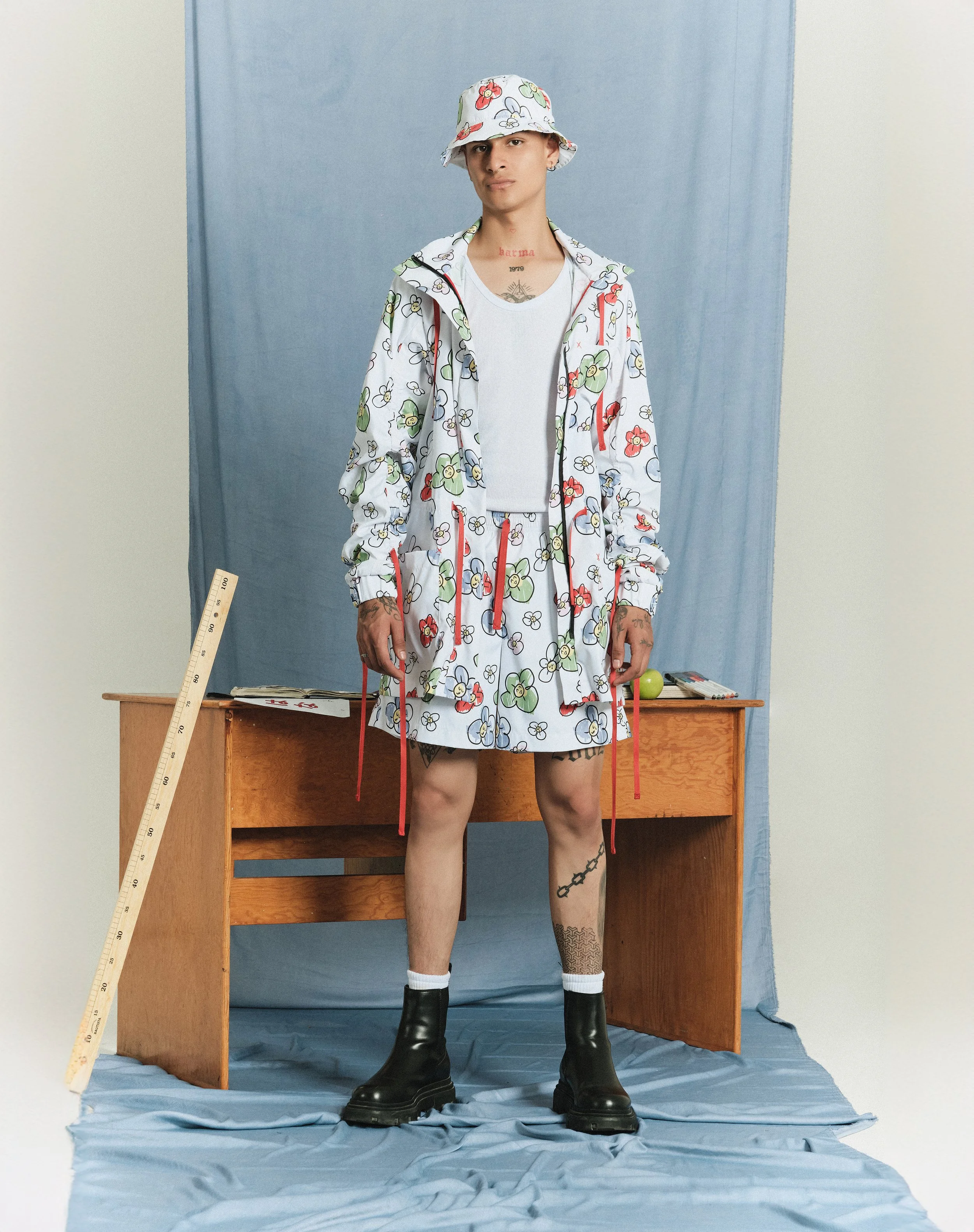 Joven con ropa estampada de flores, sombrero a juego, pantalones cortos, botas negras y calcetas blancas, posando en un fondo azul con escritorio de madera y objetos pequeños.