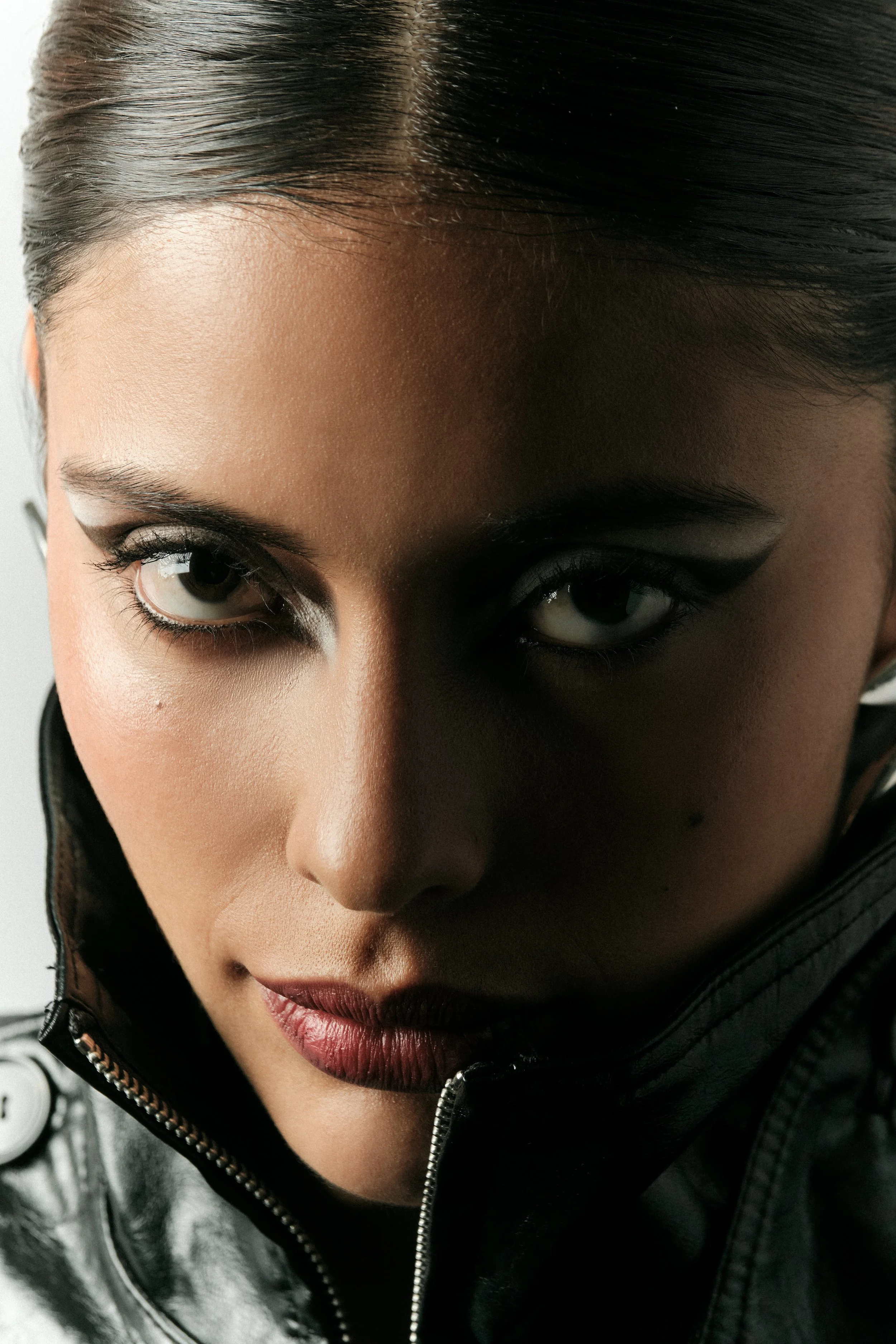 Primer plano de una mujer con maquillaje oscuro, cabello peinado hacia atrás, usando una chaqueta de cuero negra con cremallera, mirando directamente a la cámara.