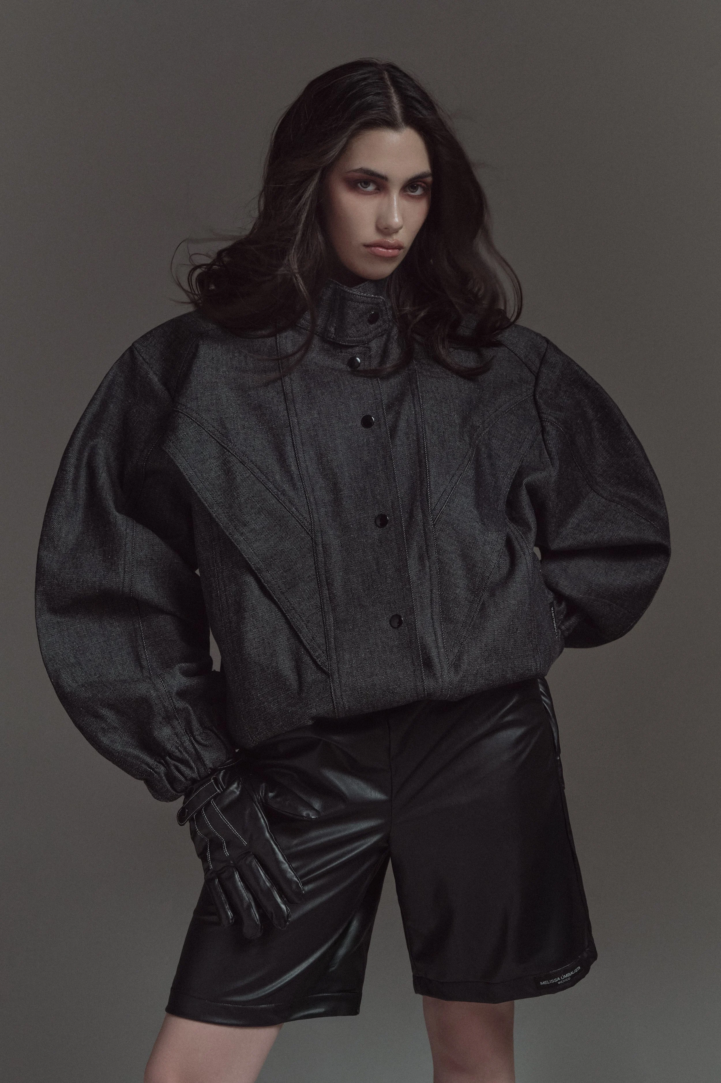 Retrato de mujer joven con cabello oscuro, vestida con chaqueta de mezclilla de color gris y pantalones negros de cuero, en fondo gris oscuro.