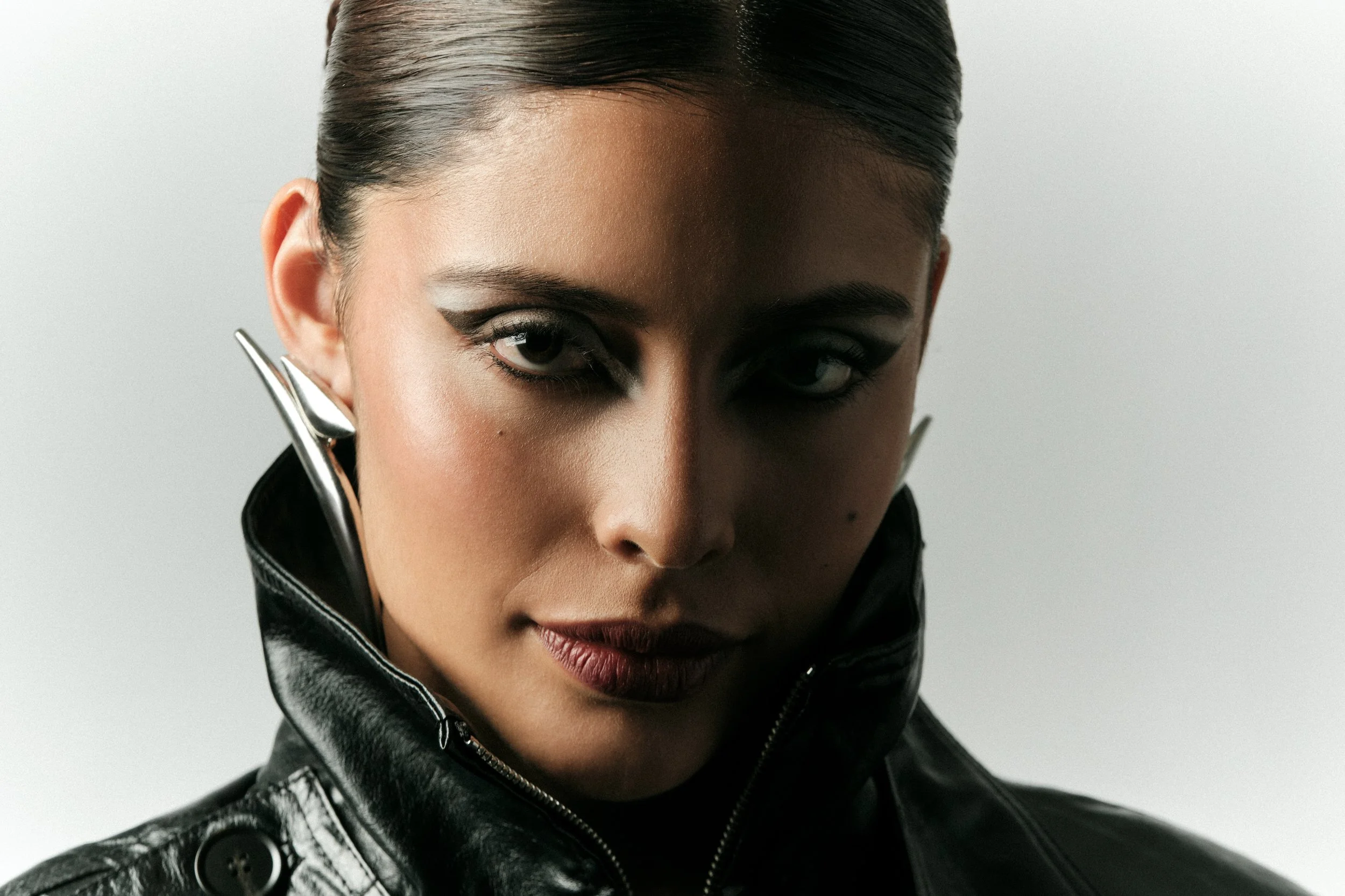Retrato de una mujer con maquillaje dramático, cabello peinado hacia atrás, usando una chaqueta de cuero negra con cierre y adornos metálicos, con expresión segura y mirada fija.