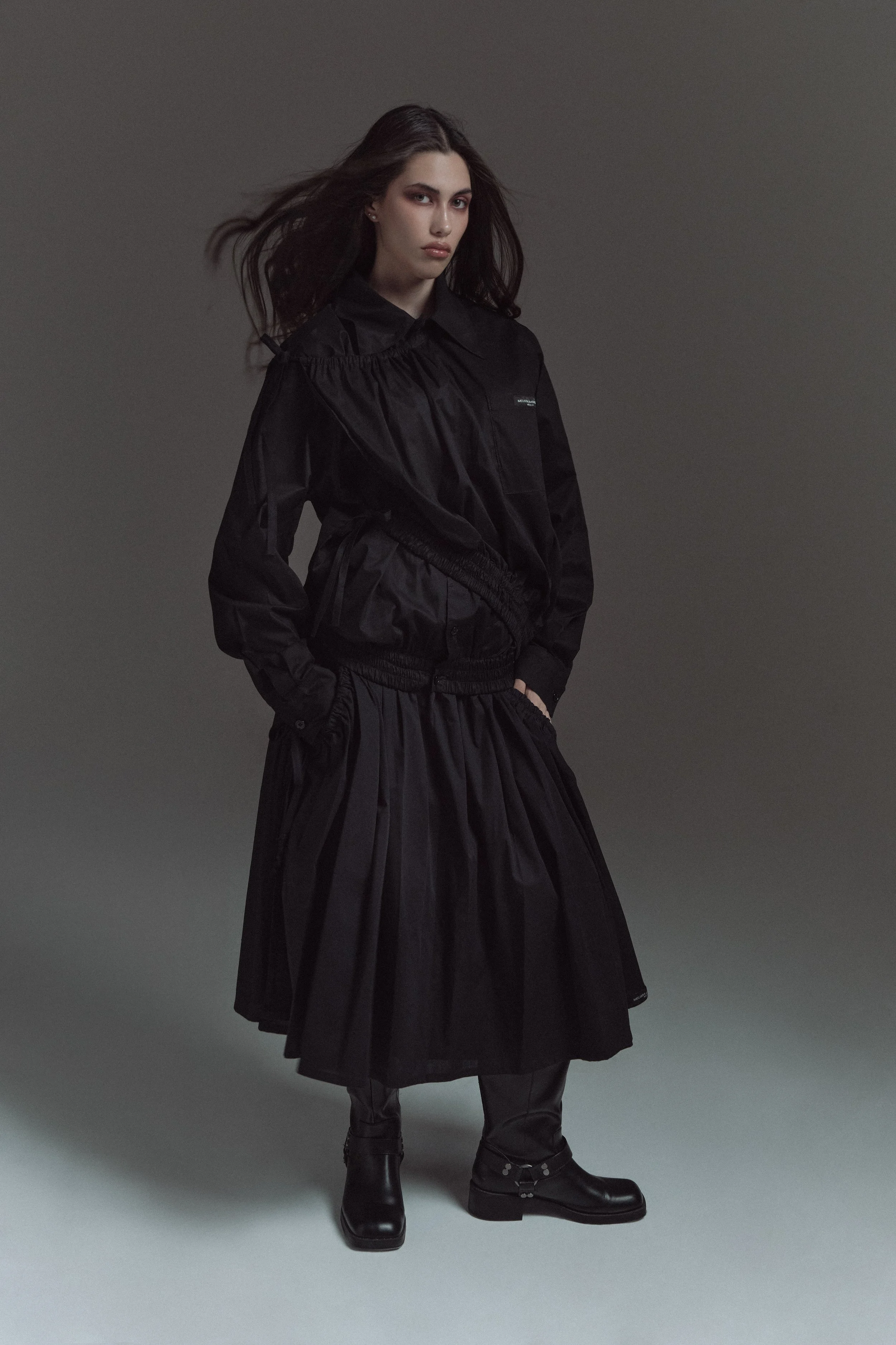 Mujer con cabello oscuro, usando chaqueta negra, falda larga negra y botas negras de cuero, de pie frente a fondo gris oscuro.