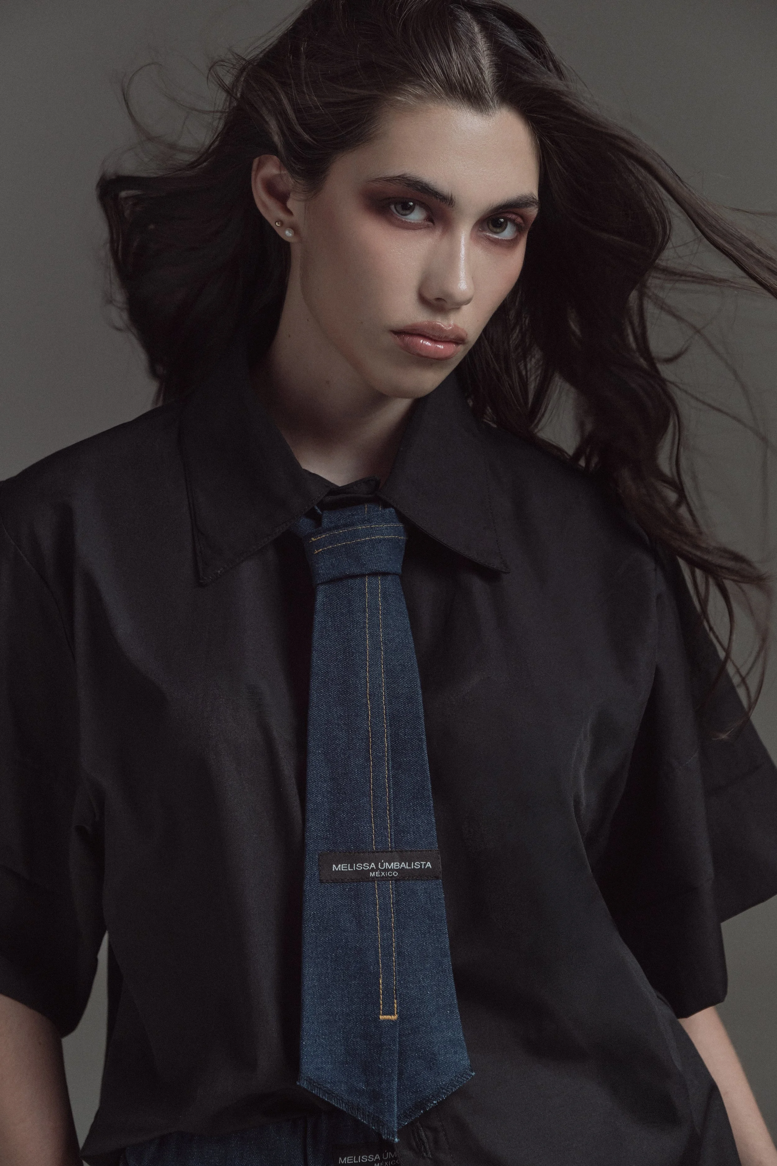 Retrato de una mujer joven con cabello largo y suelto, vestida con una camisa negra y una corbata azul con detalles de costura amarilla, en un fondo gris, con un estilo de fotografía profesional.