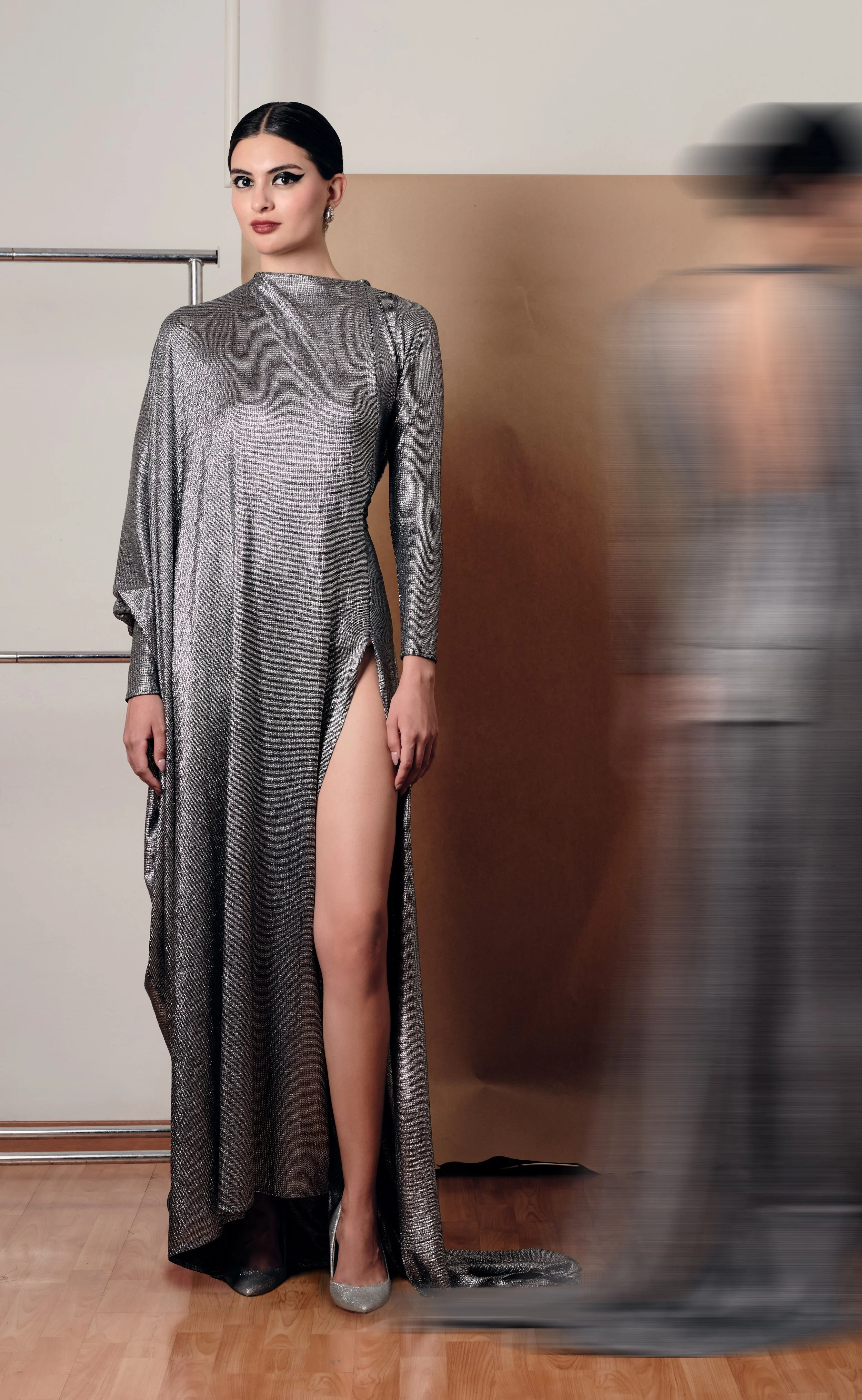 Mujer con vestido plateado de manga larga y abertura en la pierna izquierda, usando tacones plateados, en un fondo neutral con persona borrosa a la derecha.