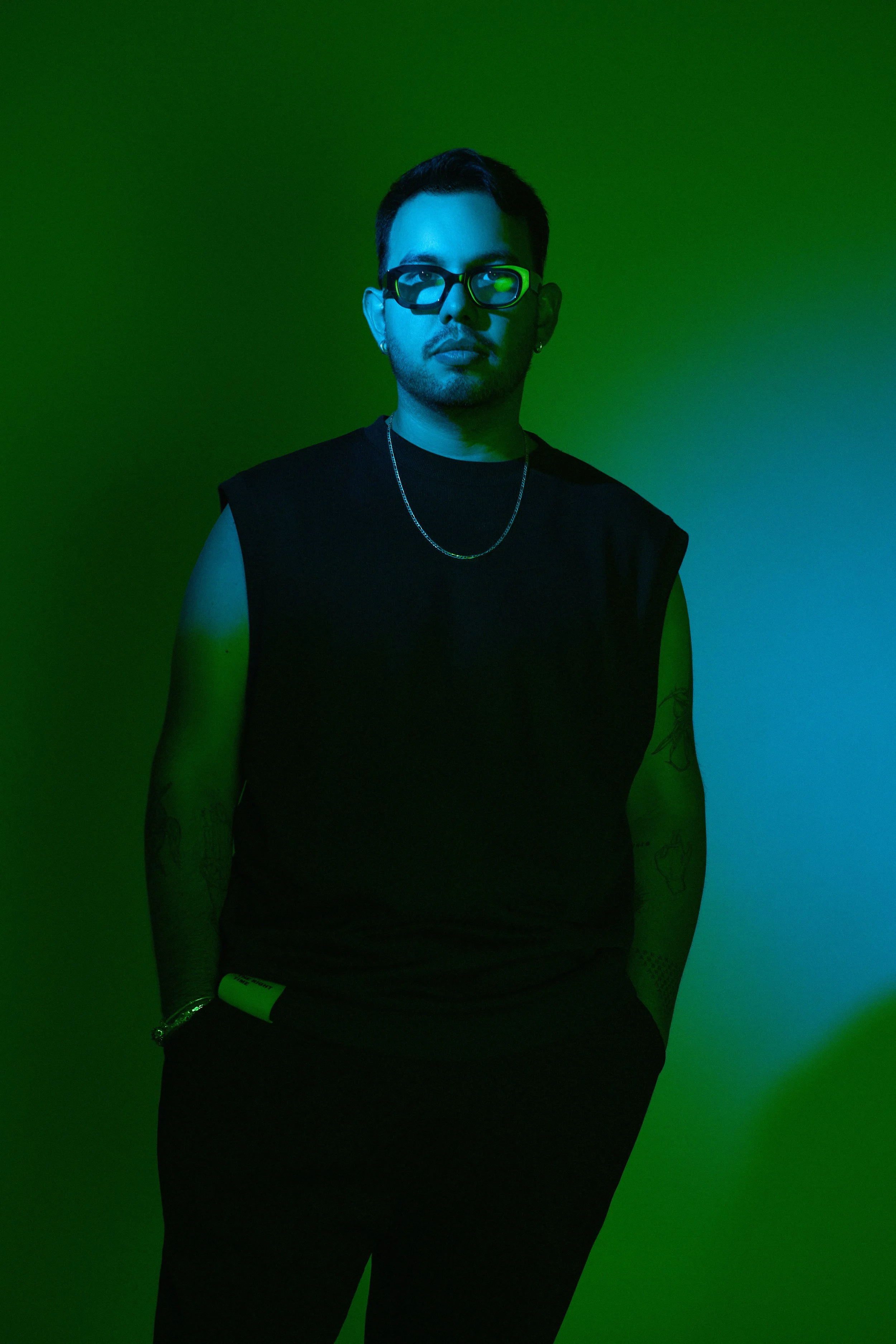 Joven con gafas, camiseta sin mangas negra, fondo en espectro de colores verdes y azules, con tatuajes en brazos, usando collar y pulsera, posando con manos en bolsillo.