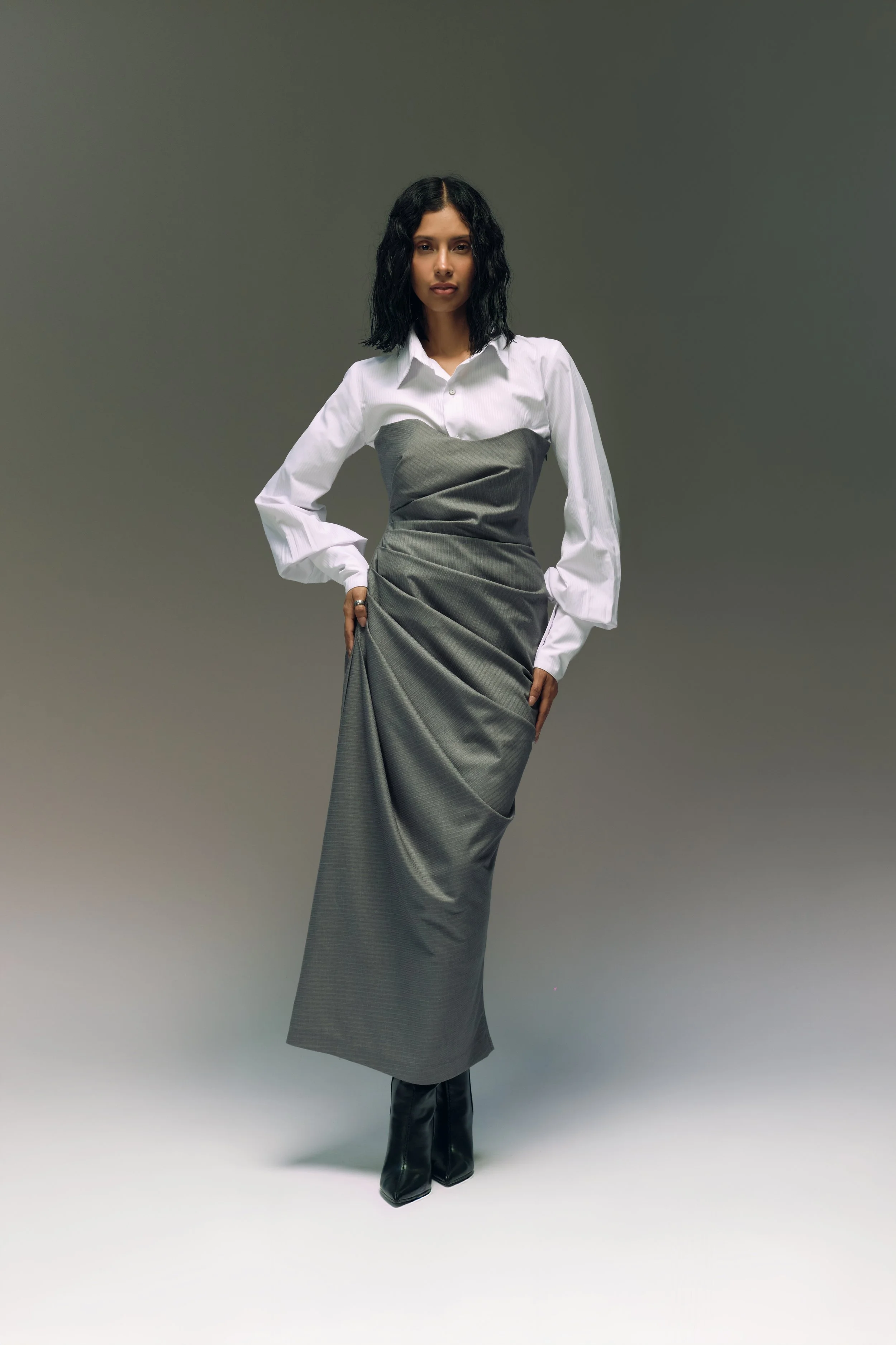 Mujer con cabello rizado negro, blusa blanca con manga larga y pantalón largo gris, posando sobre fondo gris.