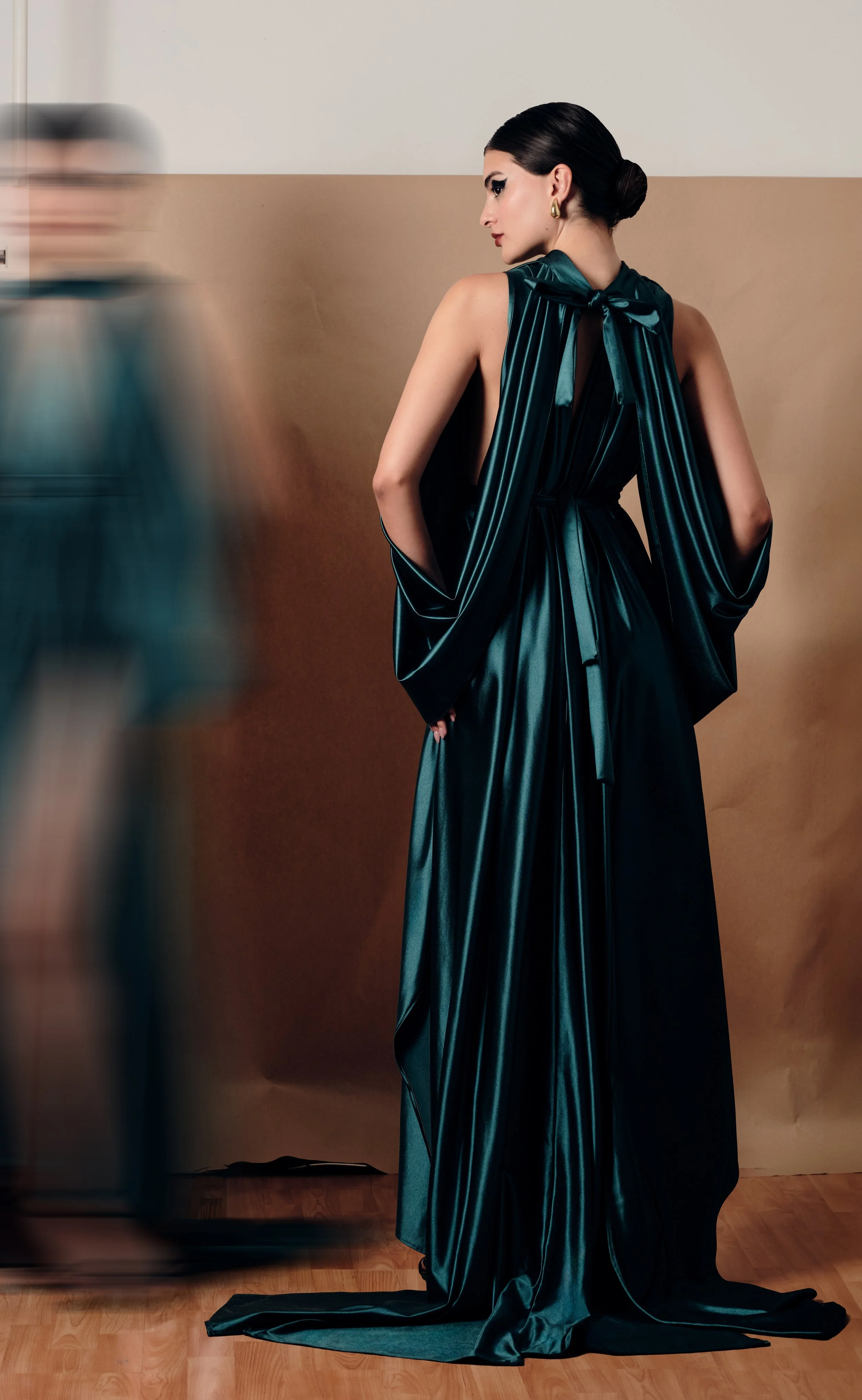 Mujer con vestido largo de satén verde oscuro y cabello peinado en chongo, posando de perfil contra pared beige.