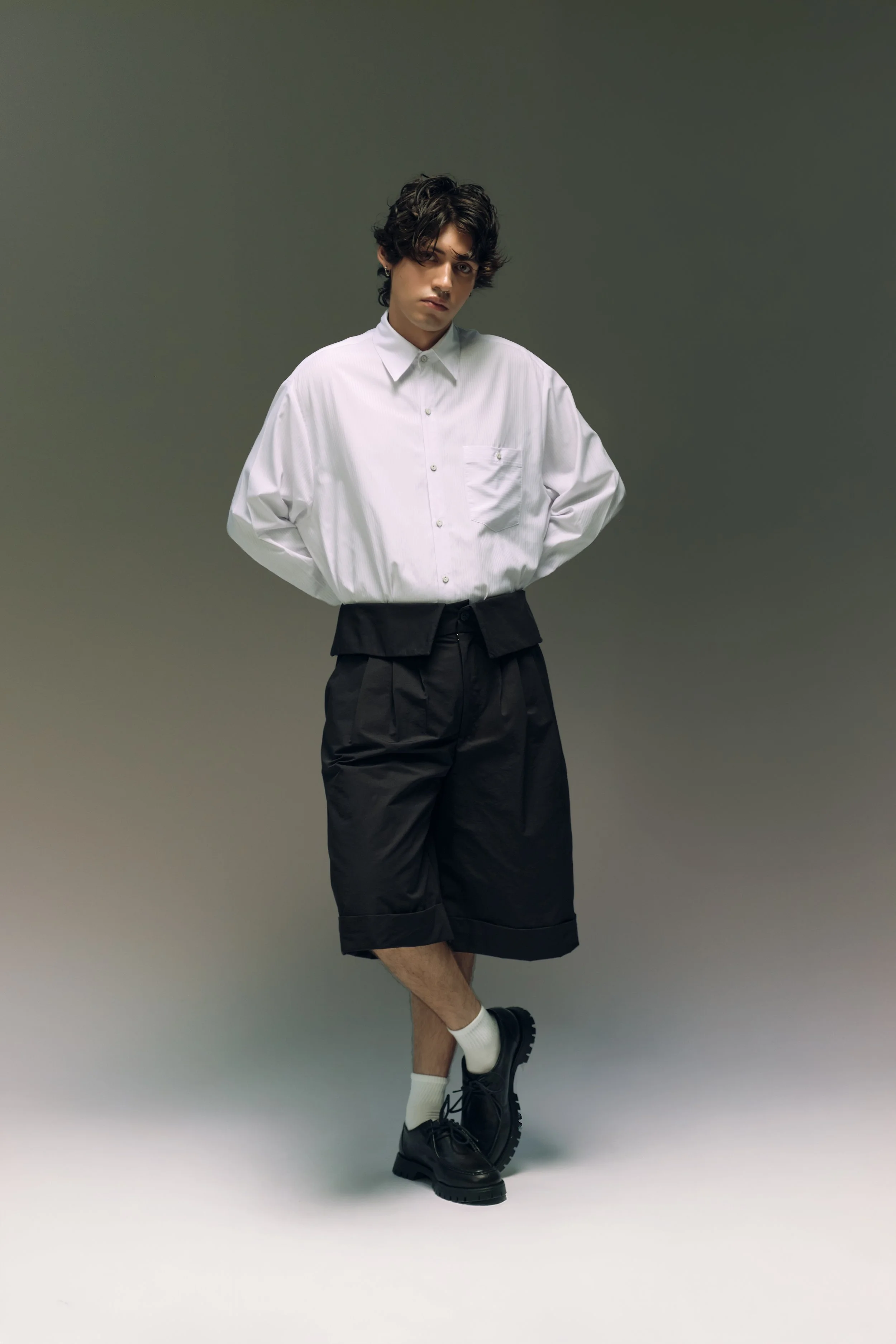 Joven con camisa blanca y pantalones cortos negros, usando botas negras y calcetas blancas, en una pose pensativa en un fondo gris.