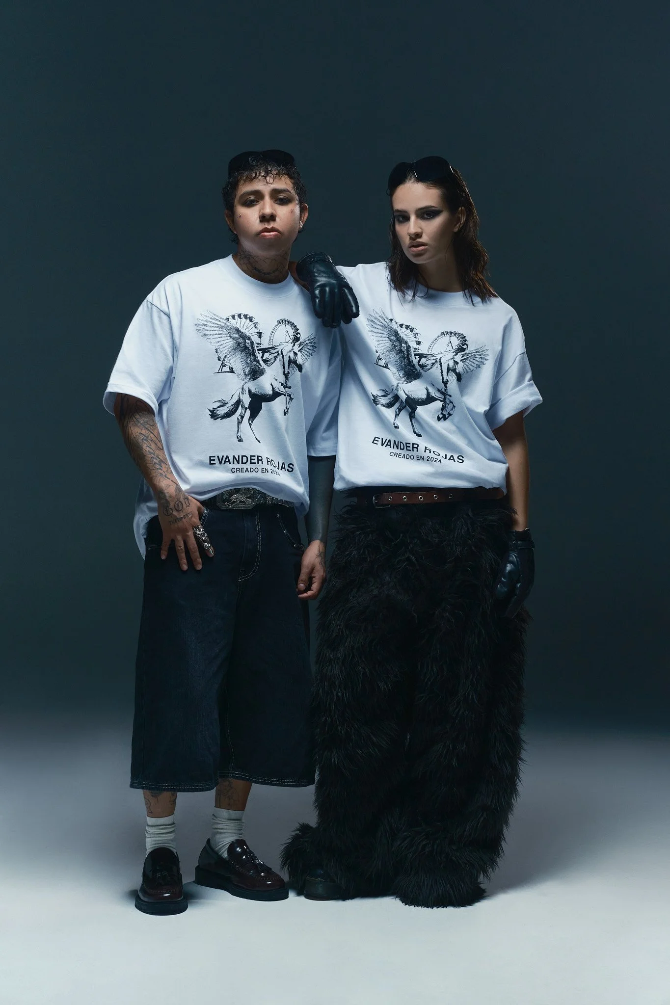 Dos personas con ropas modernas y accesorios en un fondo oscuro. Una lleva un camiseta blanca con un diseño de pegaso y la otra una camiseta similar. La persona a la izquierda usa falda larga y calzado negro con calcetas blancas; la otra lleva un pan
