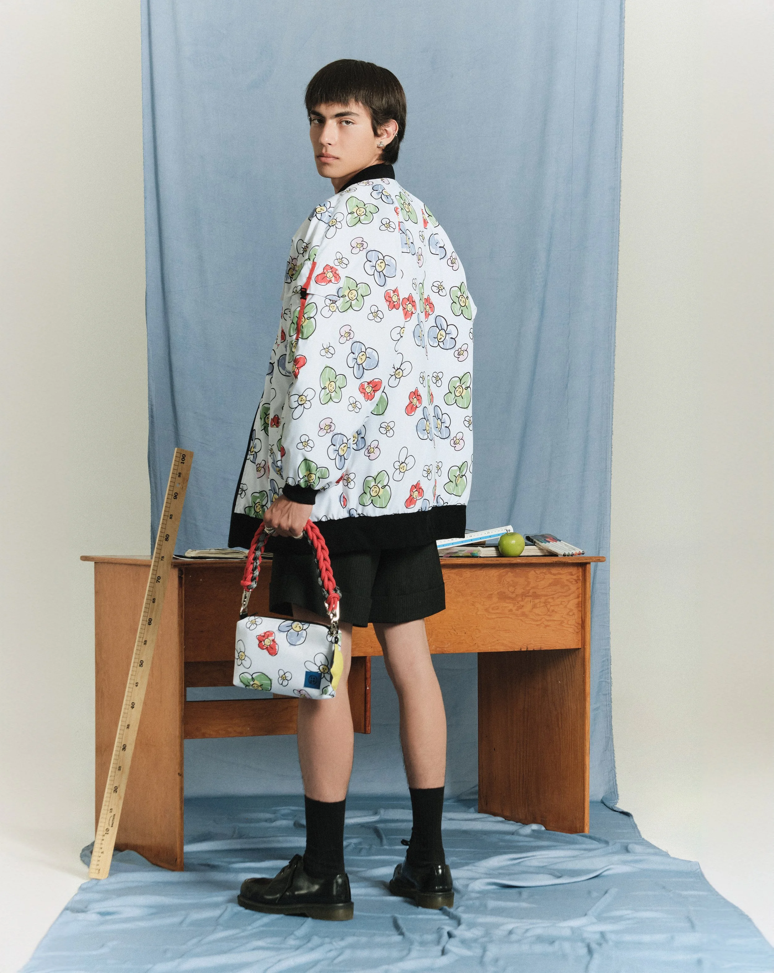 Un joven con ropa moderna y accesorios sostiene una bolsa con diseño floral, de fondo una mesa de madera con libros y una manzana, todo en un entorno con cortinas y un suelo cubierto con tela azul.