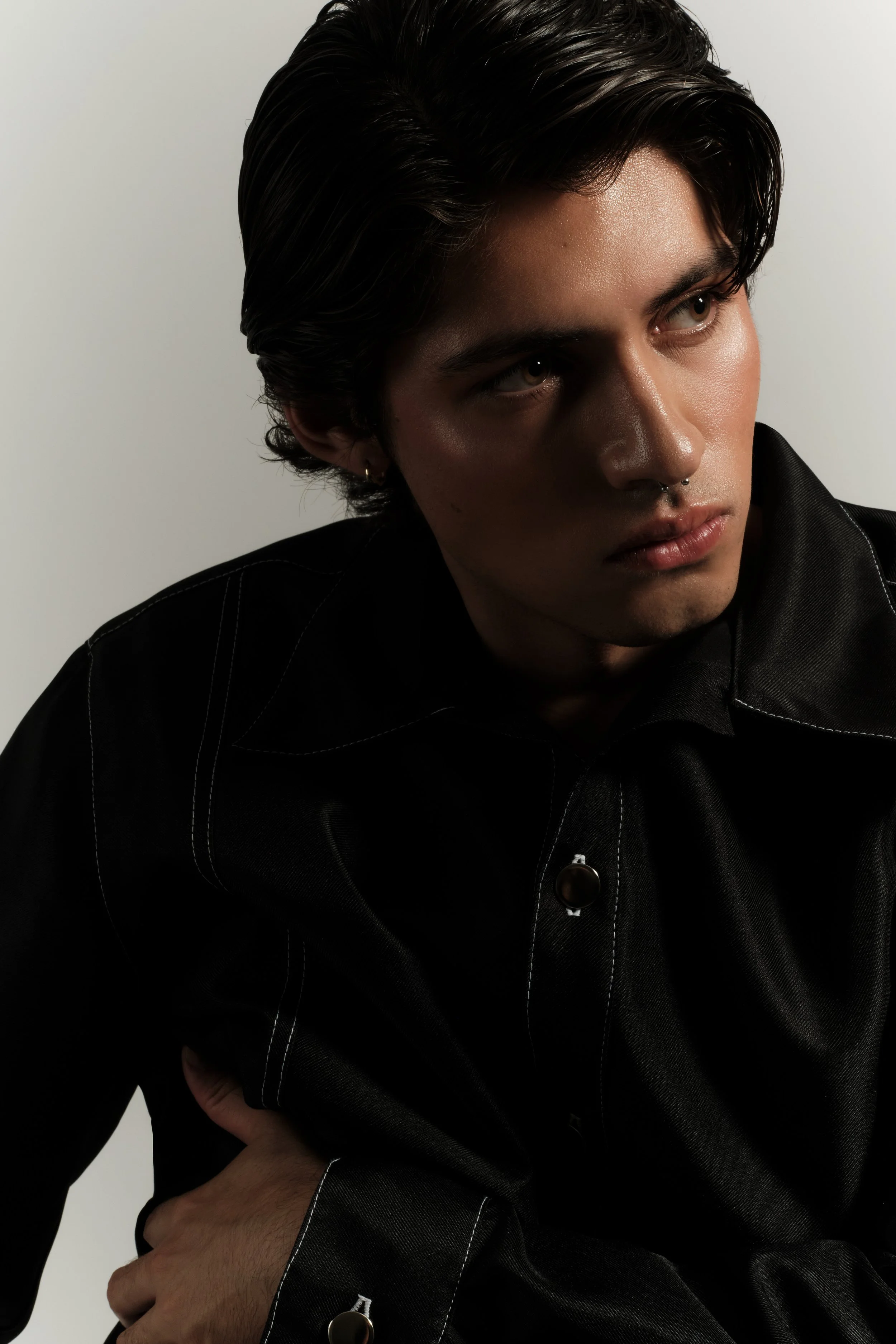 Retrato de un joven hombre con cabello oscuro y peinado hacia atrás, usando una camisa negra con costuras blancas, posando en un fondo blanco y gris.