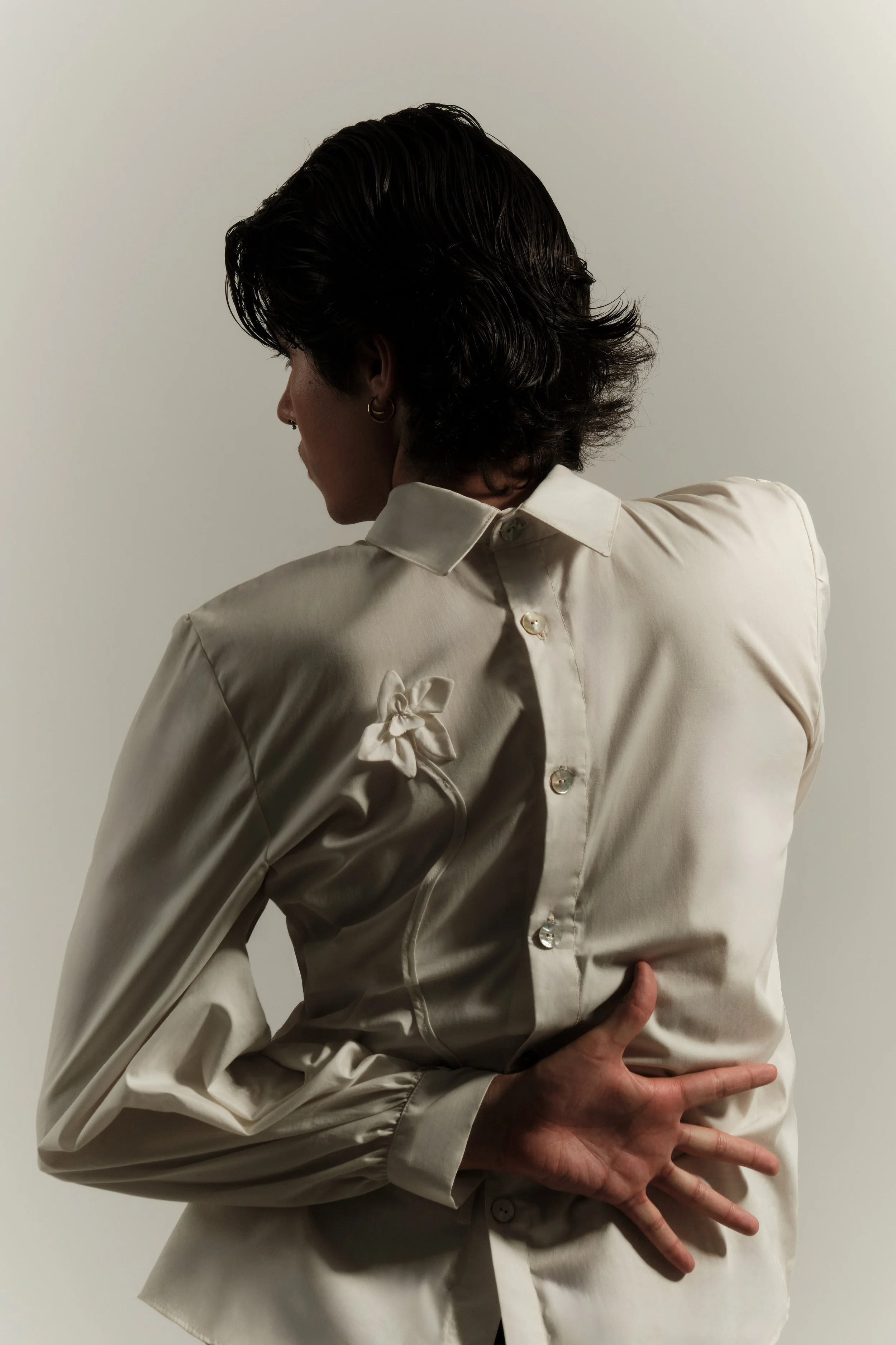 Mujer con cabello oscuro, vistiendo una blusa de seda beige con un lazo en la espalda, tocando la parte baja de su espalda con la mano izquierda, en un fondo gris claro.