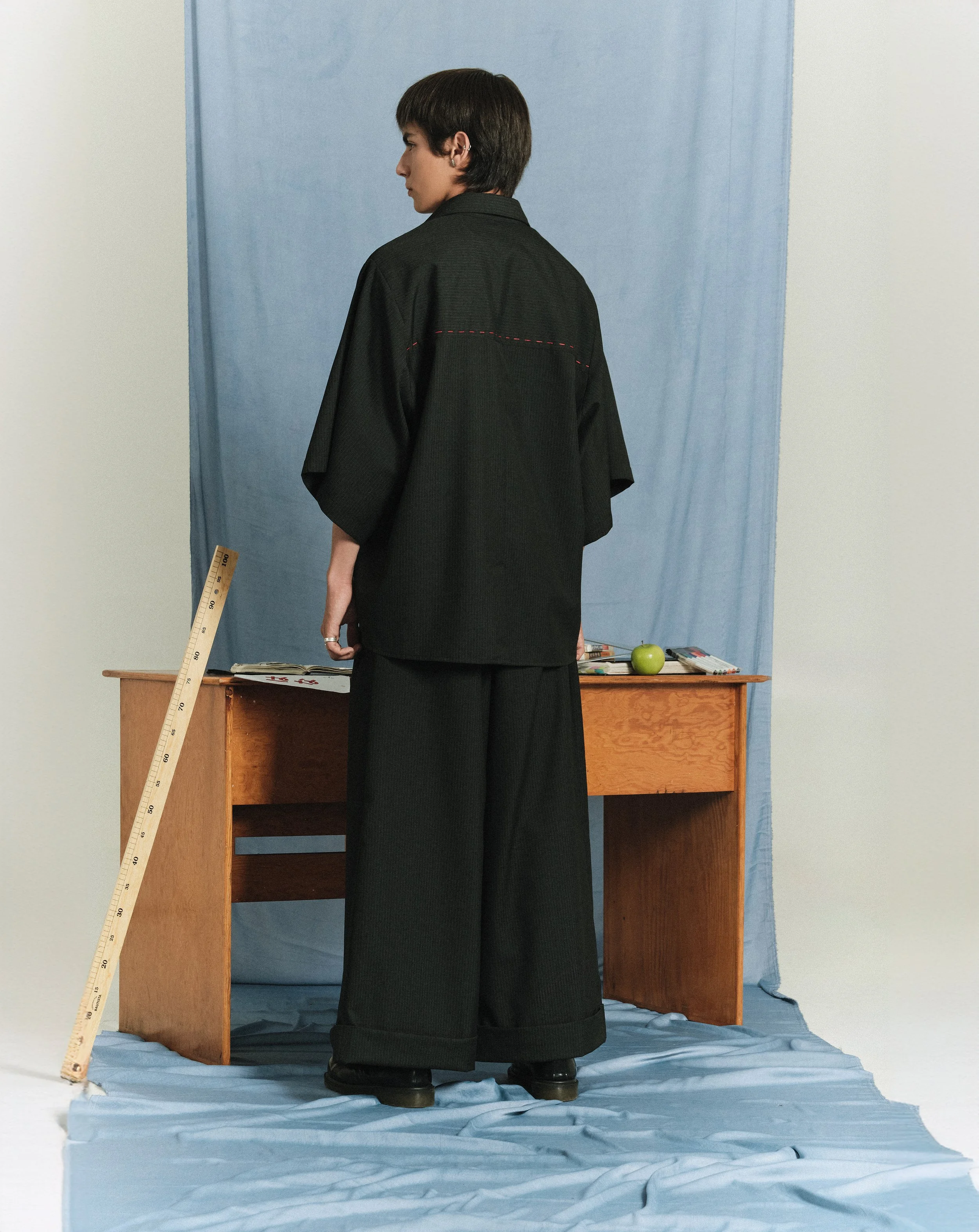 Persona con cabello castaño oscuro y ropa negra, de espaldas, de pie frente a un escritorio con objetos y un fondo azul, en una escena de estudio.