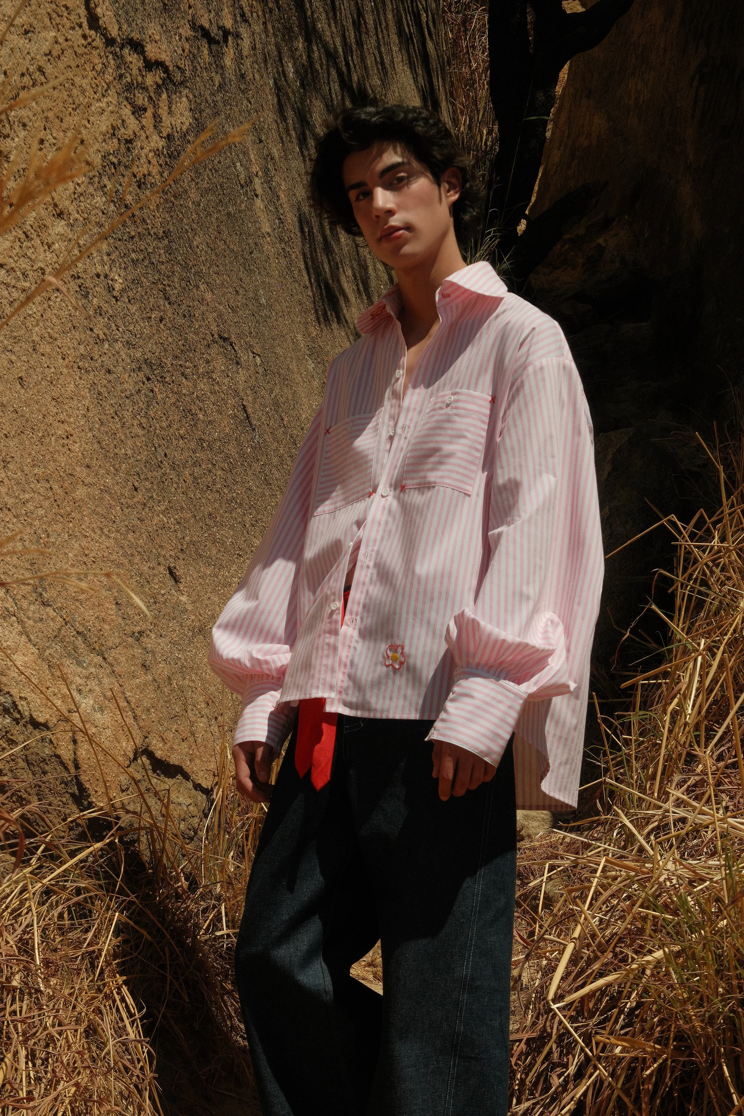 Joven con camisa de rayas rosa y blanca y pantalones oscuros en un paisaje al aire libre con rocas y vegetación seca.