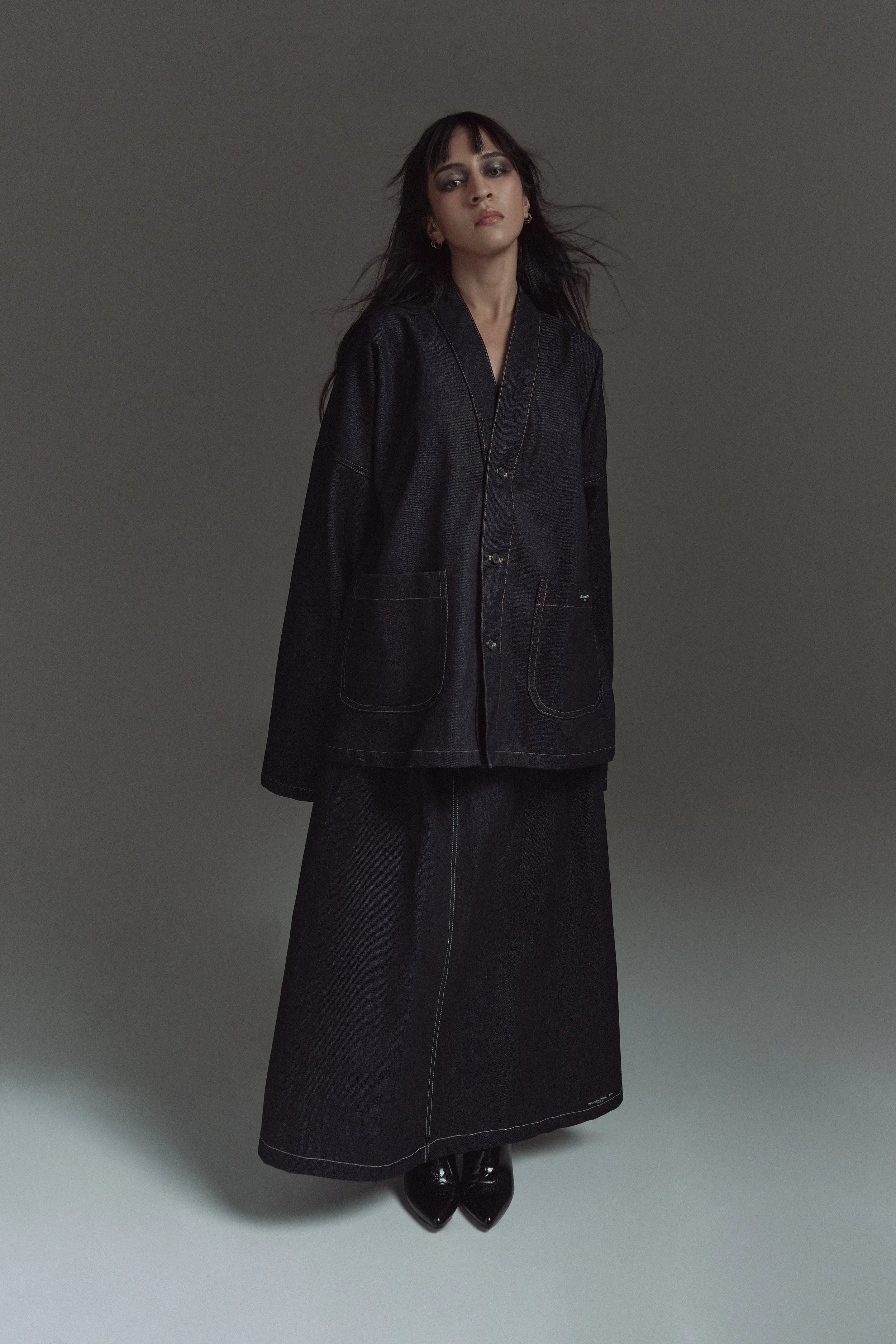 Mujer con cabello oscuro y maquillaje, vestida con un conjunto de mezclilla oscuro y botas negras, posando de pie contra un fondo gris
