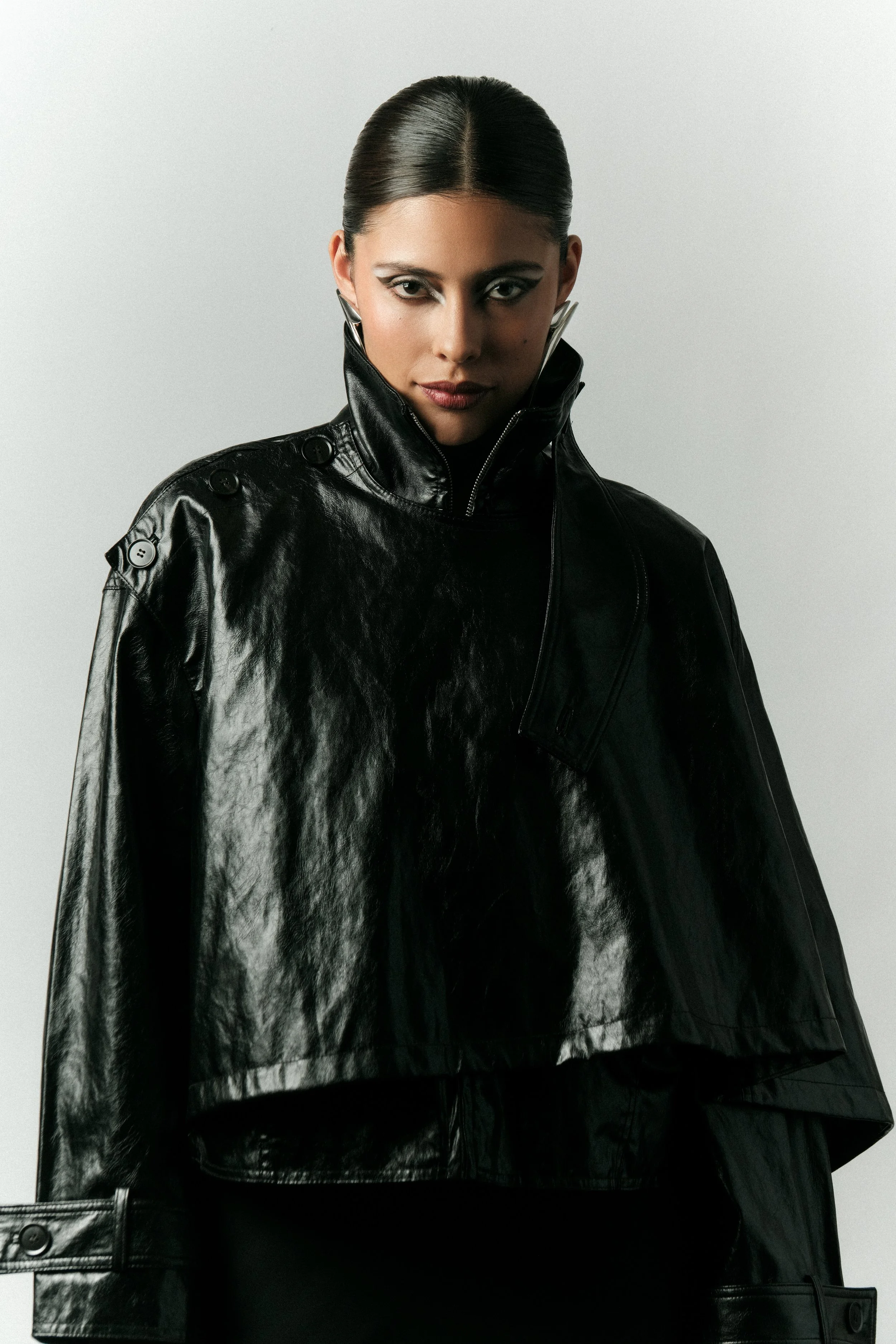 Mujer con cabello recogido, vestida con chaqueta de cuero negra, mirando intensamente, fondo gris claro.