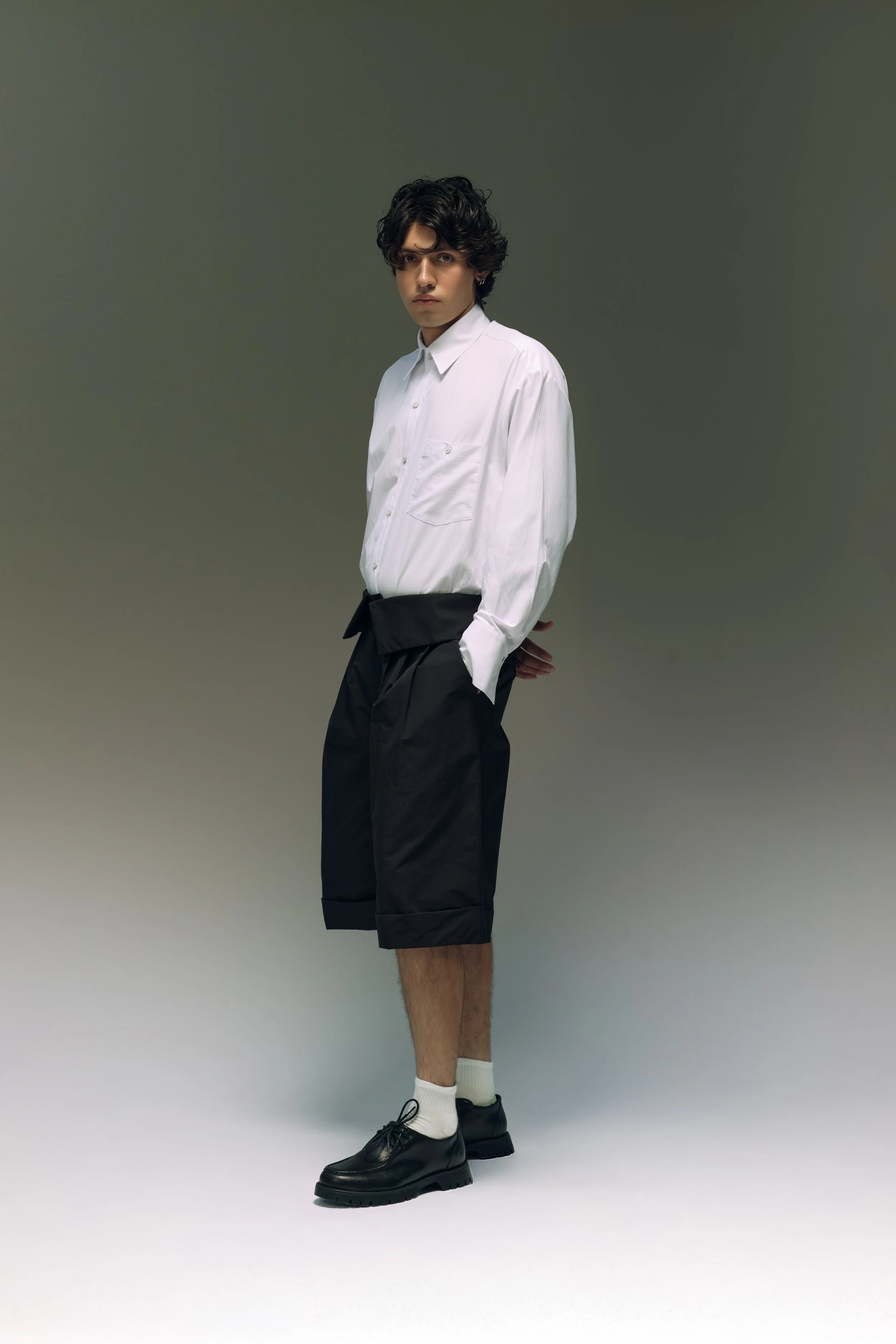 Joven hombre con cabello oscuro, vistiendo camisa blanca y pantalones cortos negros, con zapatos negros y calcetines blancos, en fondo gris.