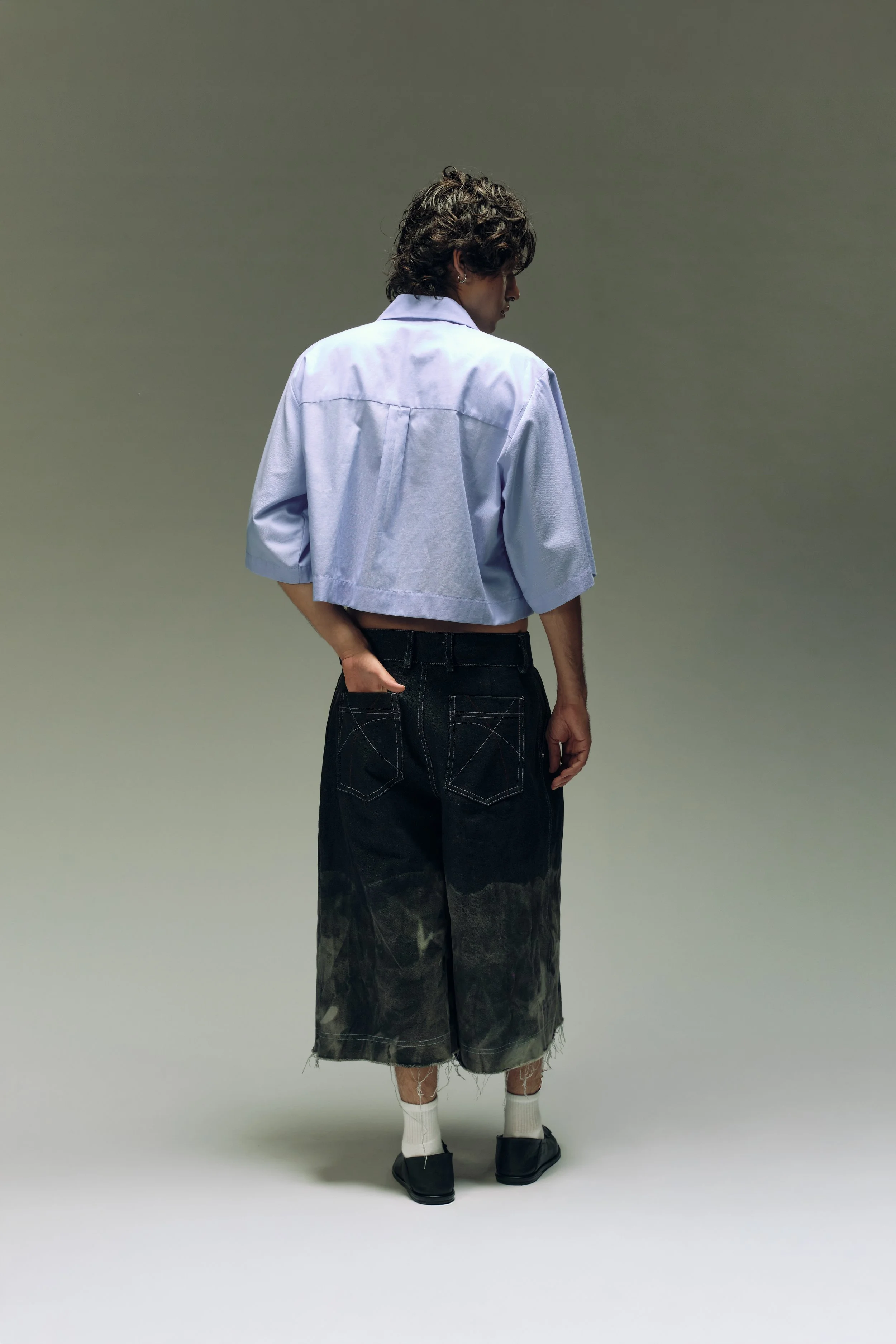 Persona de espaldas con camiseta blanca cortada, pantalones a la moda y calcetas blancas, en un fondo gris liso.