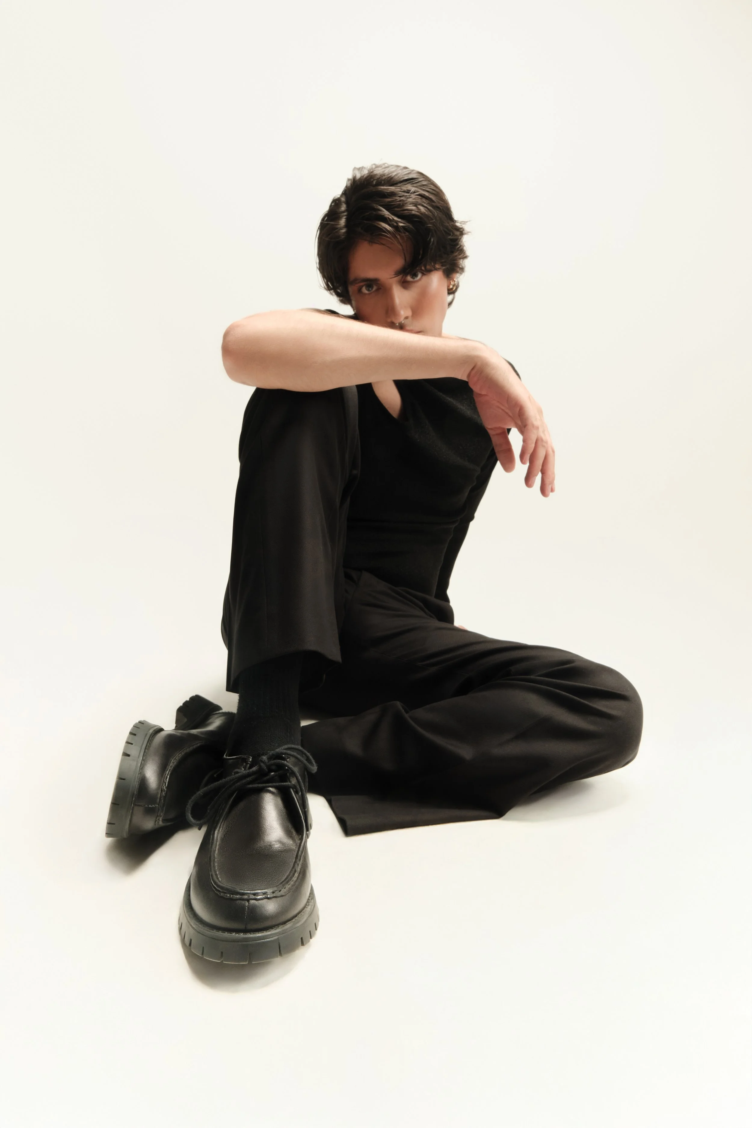 Joven con ropa negra sentado en el suelo con pierna cruzada, en un fondo blanco.