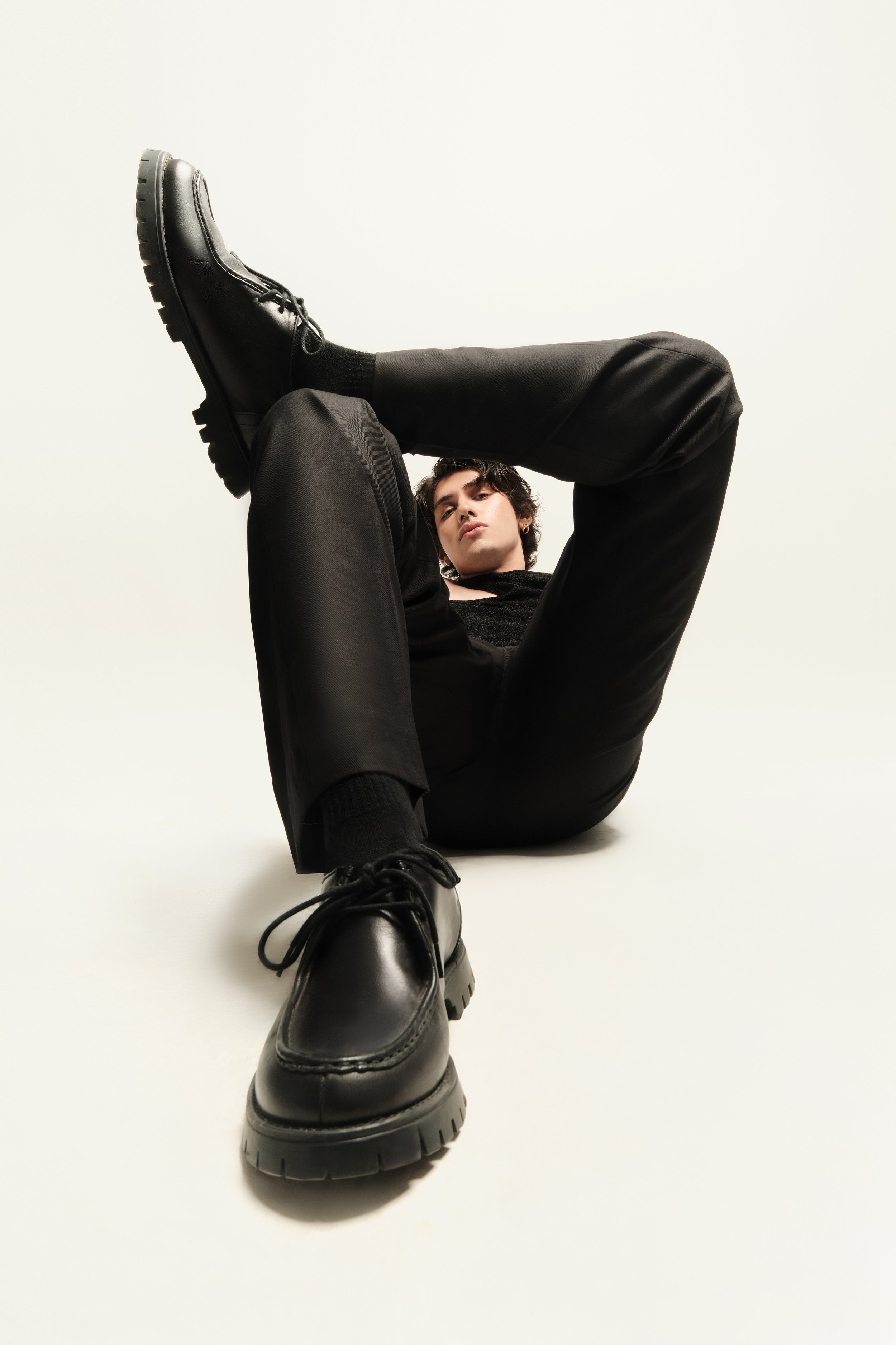 Persona joven sentada en el suelo con una pierna cruzada sobre la otra, vestida con ropa negra y botas negras de suela gruesa, mirada hacia la cámara, fondo blanco.