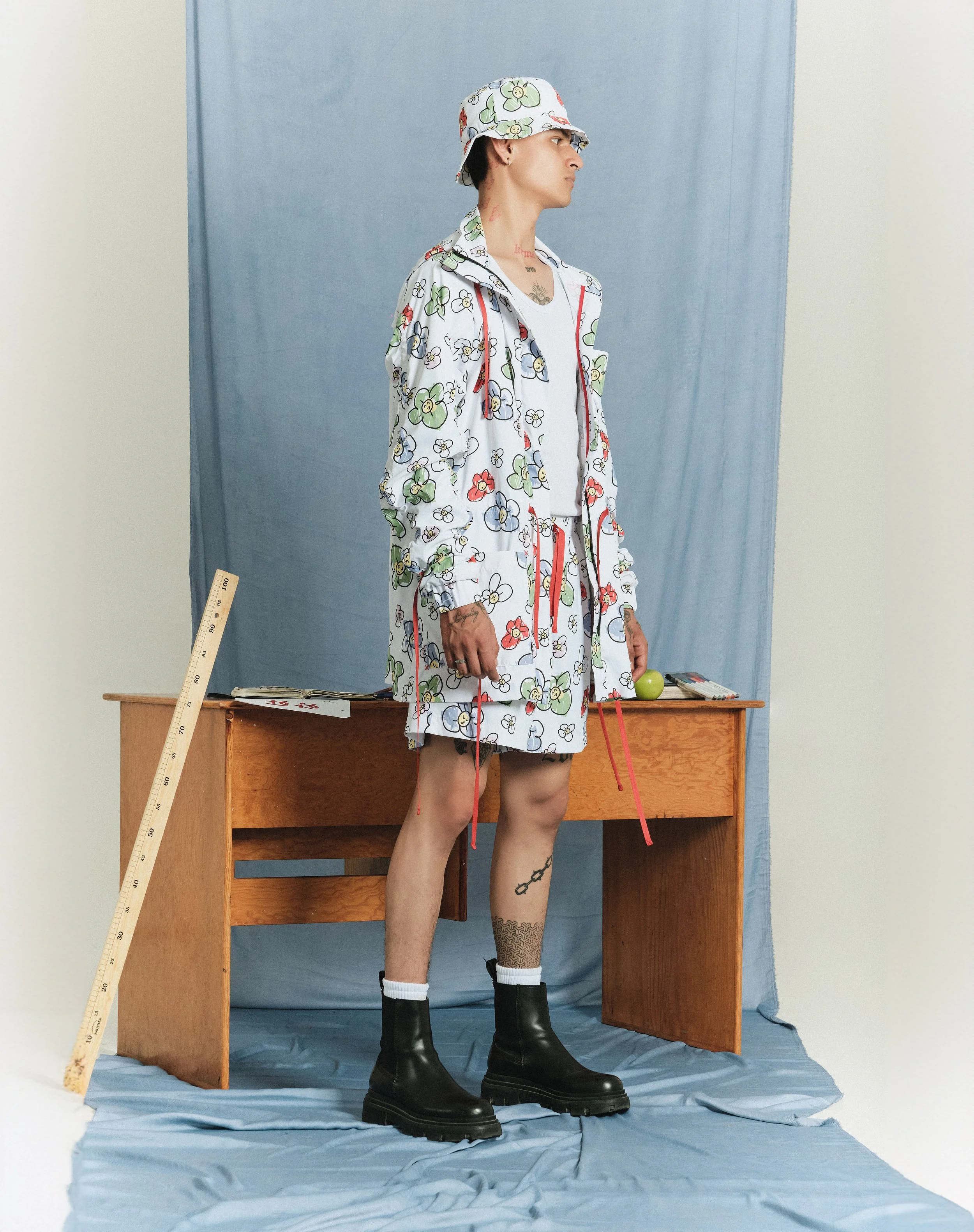 Joven con ropa decorada con flores, sombrero, botas negras, de pie junto a un escritorio en un fondo de tela azul.