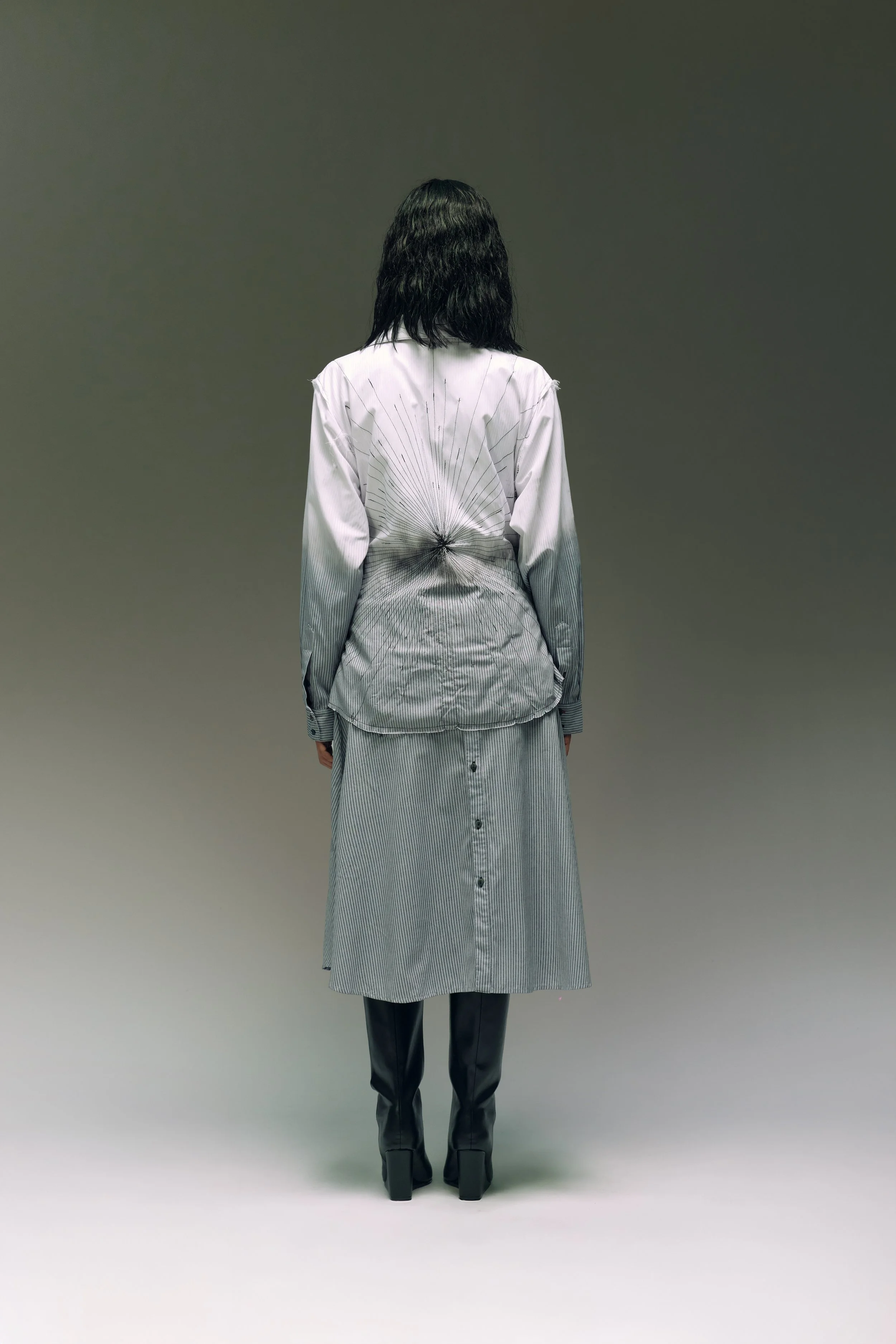 Persona de espaldas, usando ropa con diseño de líneas finas y en tonos grises, en un fondo simple y gris.