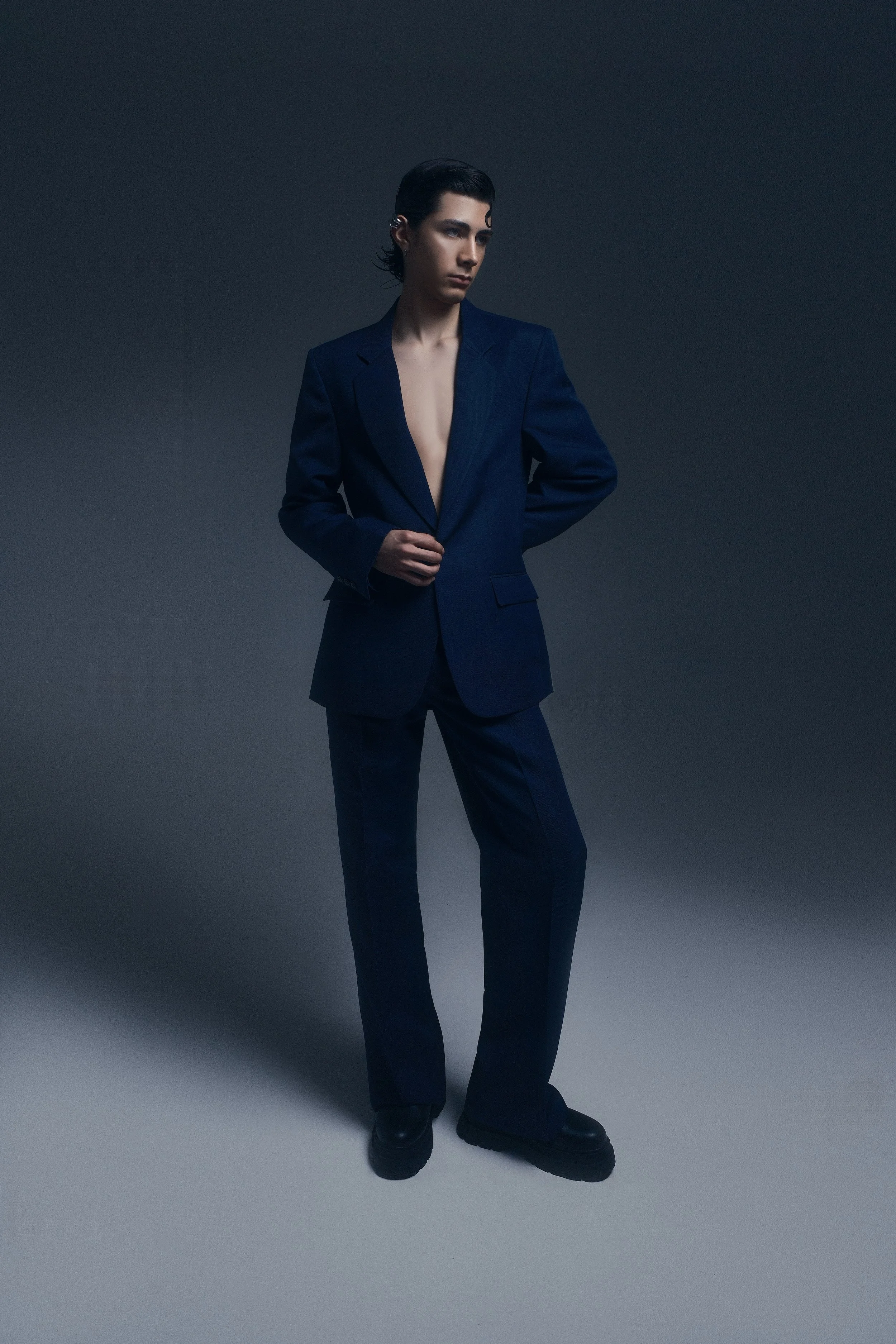 Joven con cabello oscuro, vestido con traje azul, de pie contra un fondo gris oscuro.