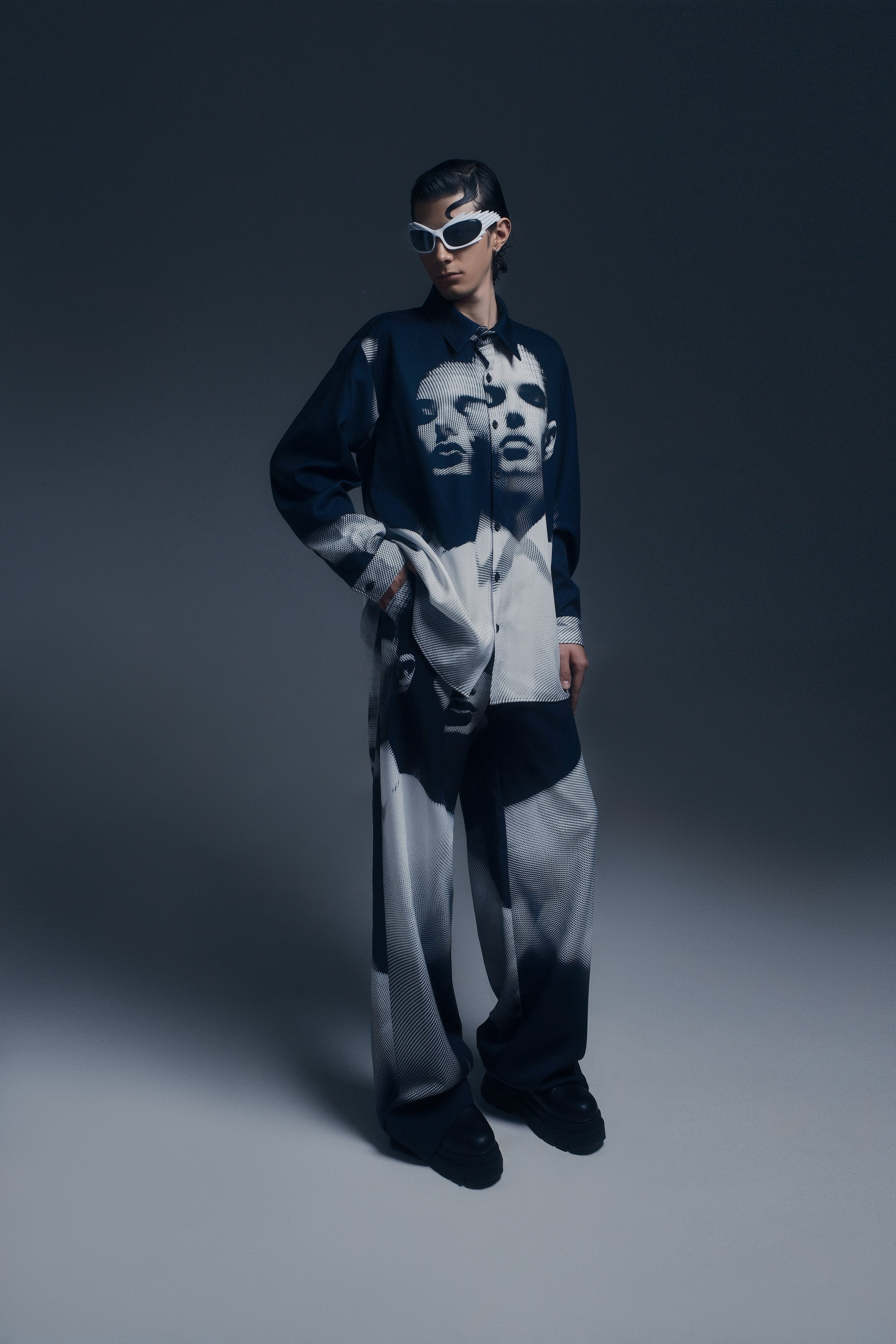 Persona con ropa moderna y gafas grandes en fondo gris oscuro.