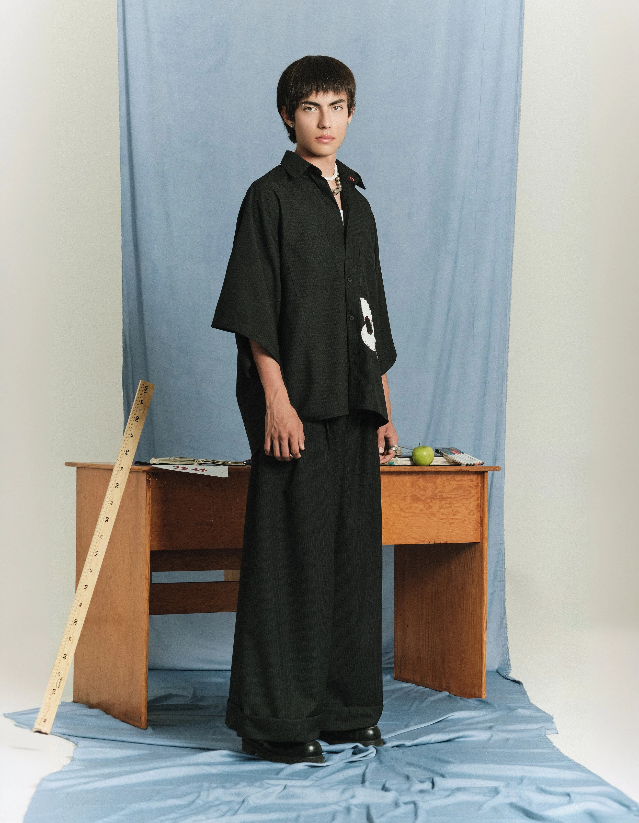 Persona de pie frente a un fondo azul, usando ropa negra y una cadena, junto a un escritorio con una manzana y otros objetos, y una regla apoyada en la mesa.