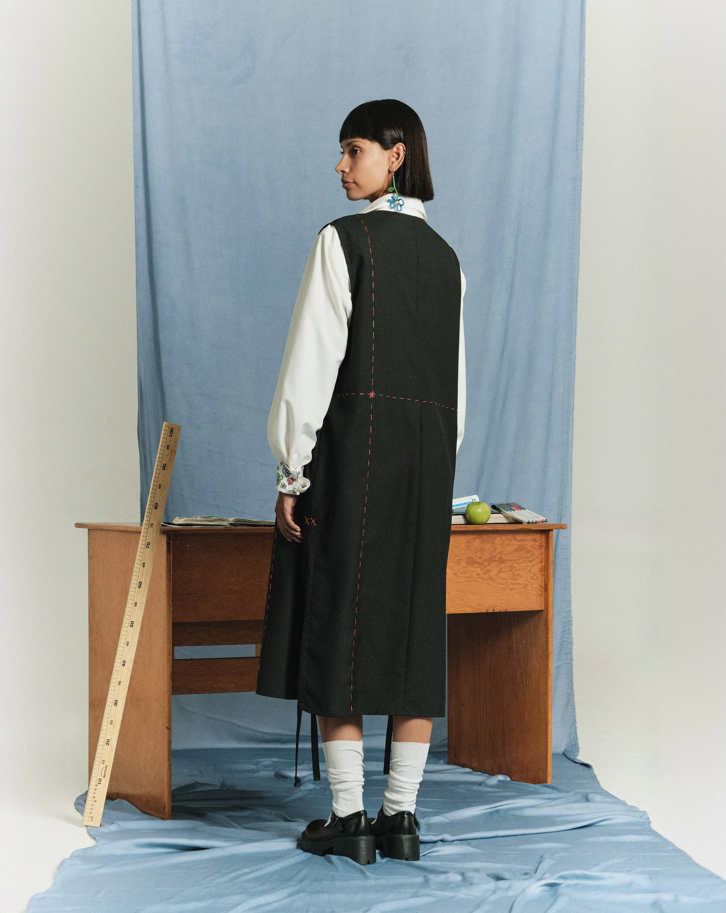 Mujer con cabello corto y oscuro, vestida con una camisa blanca, un vestido negro largo y zapatos con calcetas blancas, de pie frente a un escritorio de madera en un ambiente con telón azul y fondo gris, con regla de madera apoyada a la izquierda.