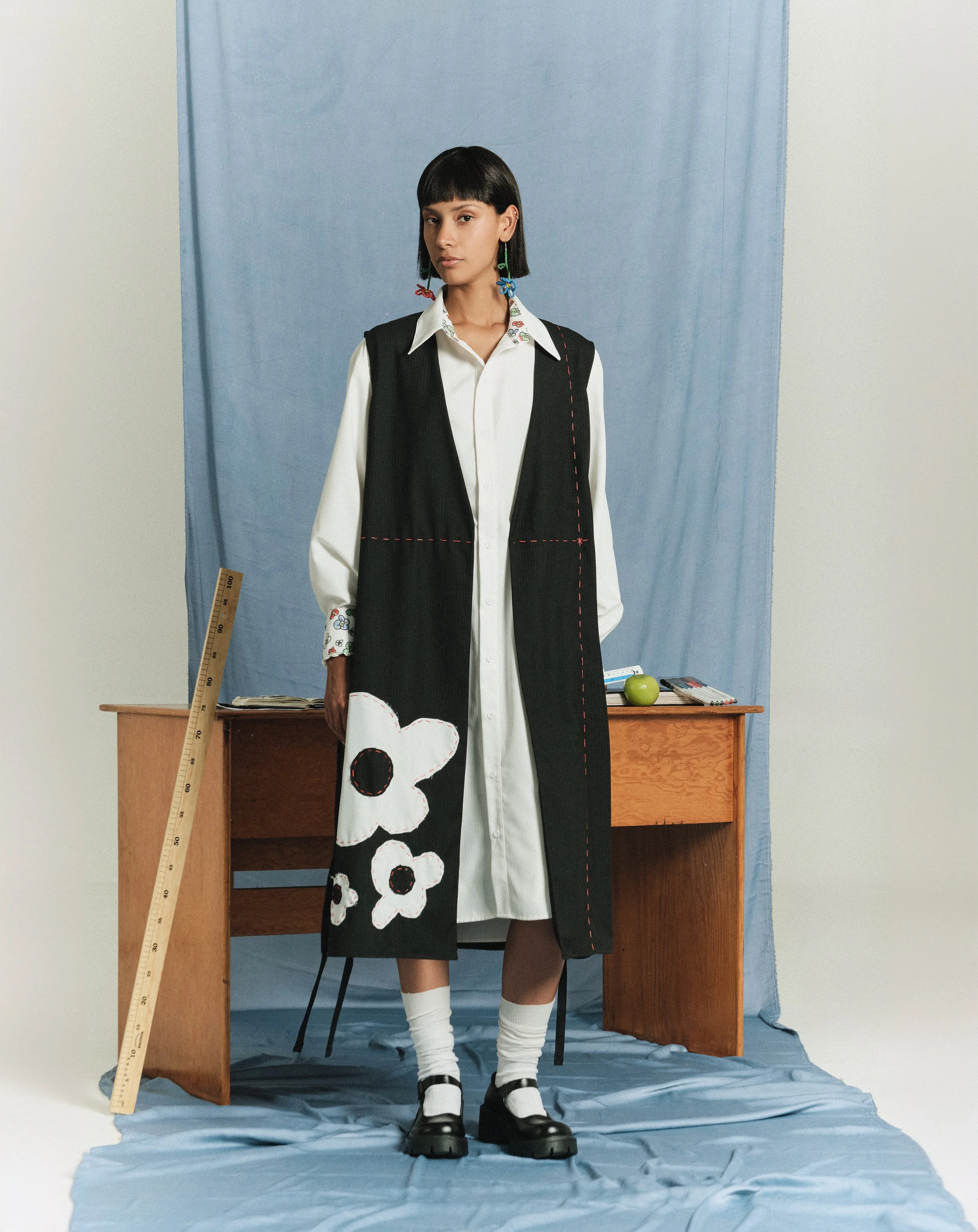 Joven usando vestido largo negro con bordados de flores blancas y detalles de costura roja, camisa blanca con bordados en las mangas, en un entorno con fondo azul y escritorio de madera con objetos de oficina.