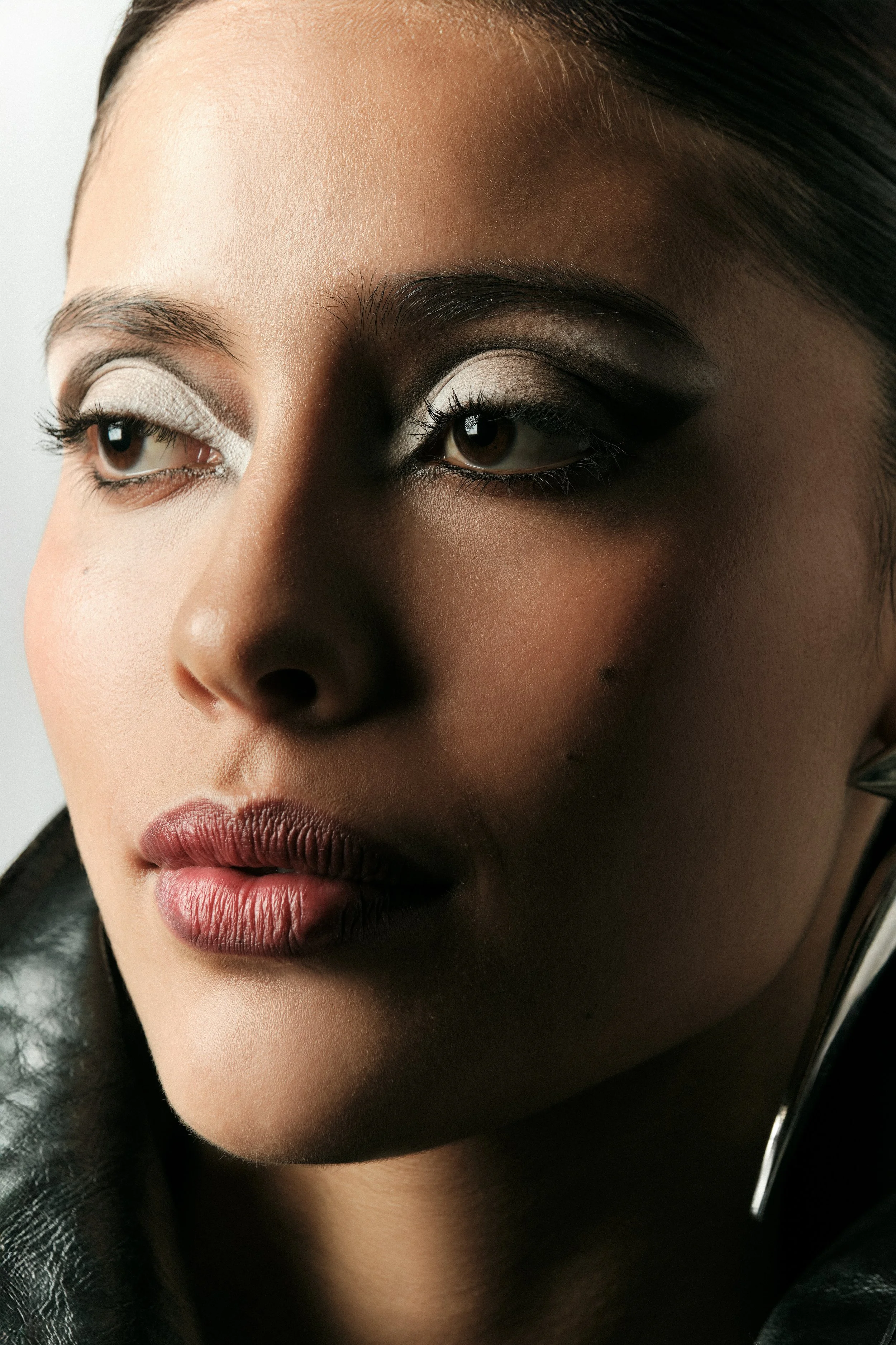 Primer plano de una mujer con maquillaje artístico, ojos con sombra de ojos blanca y negra, labios con lápiz labial nude, piel suave y cabello oscuro peinado hacia atrás.