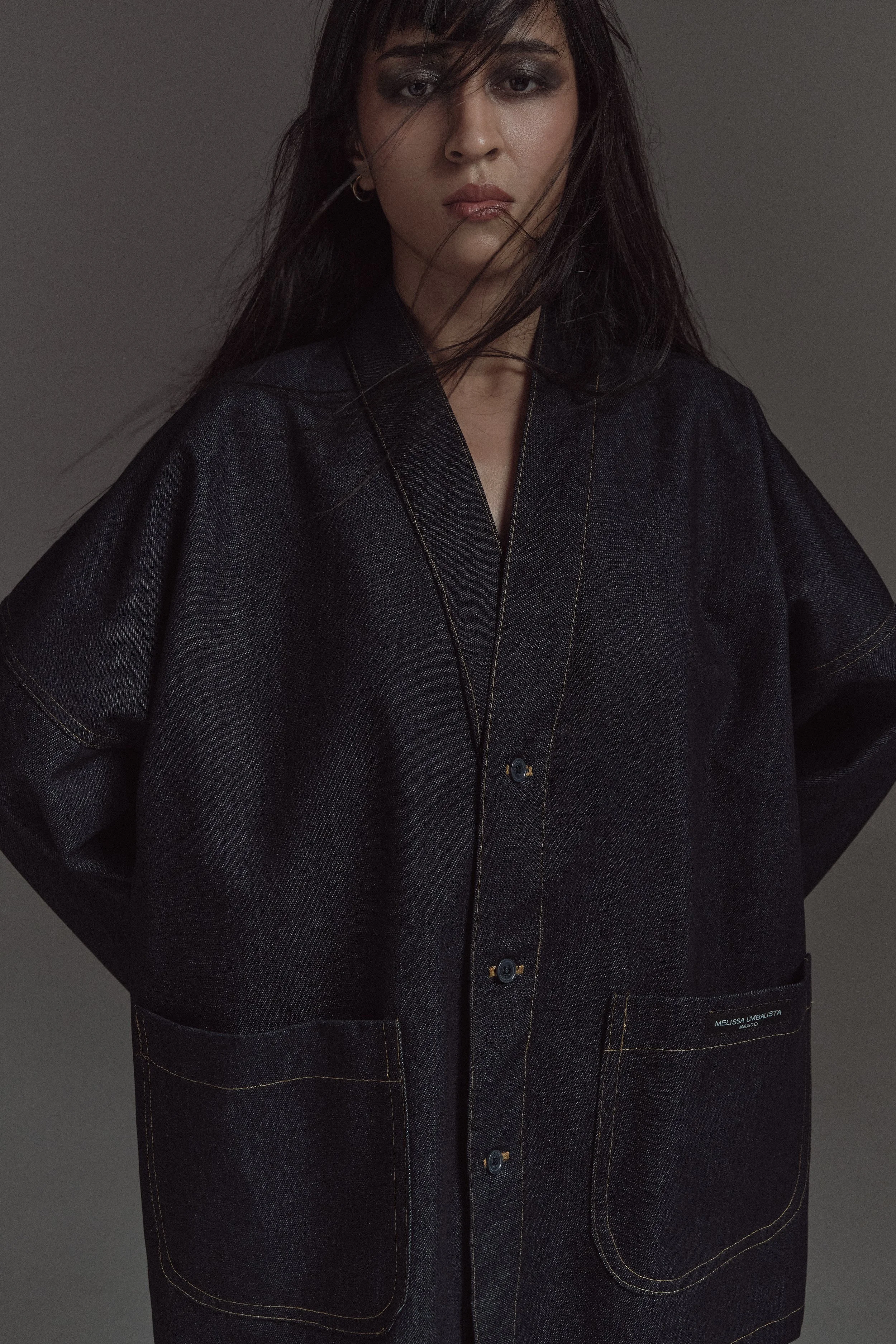 Mujer con pelo largo y oscuro, lleva una blusa o vestido negro con costuras en contraste y botones, en un fondo gris oscuro.