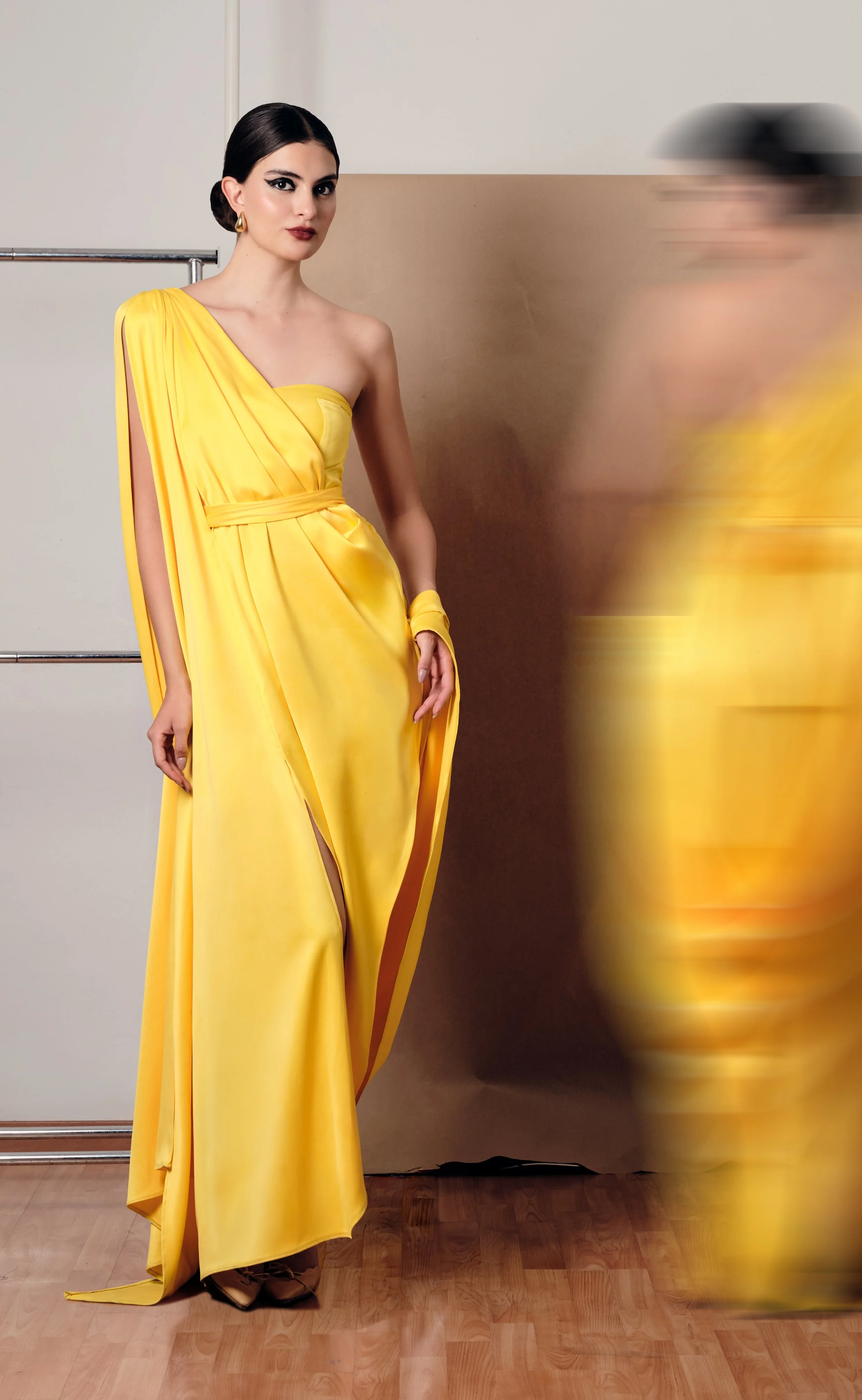 Mujer con vestido amarillo largo y peinado recogido, de pie junto a fondo beige y pared blanca, con movimiento borroso en su derecha.