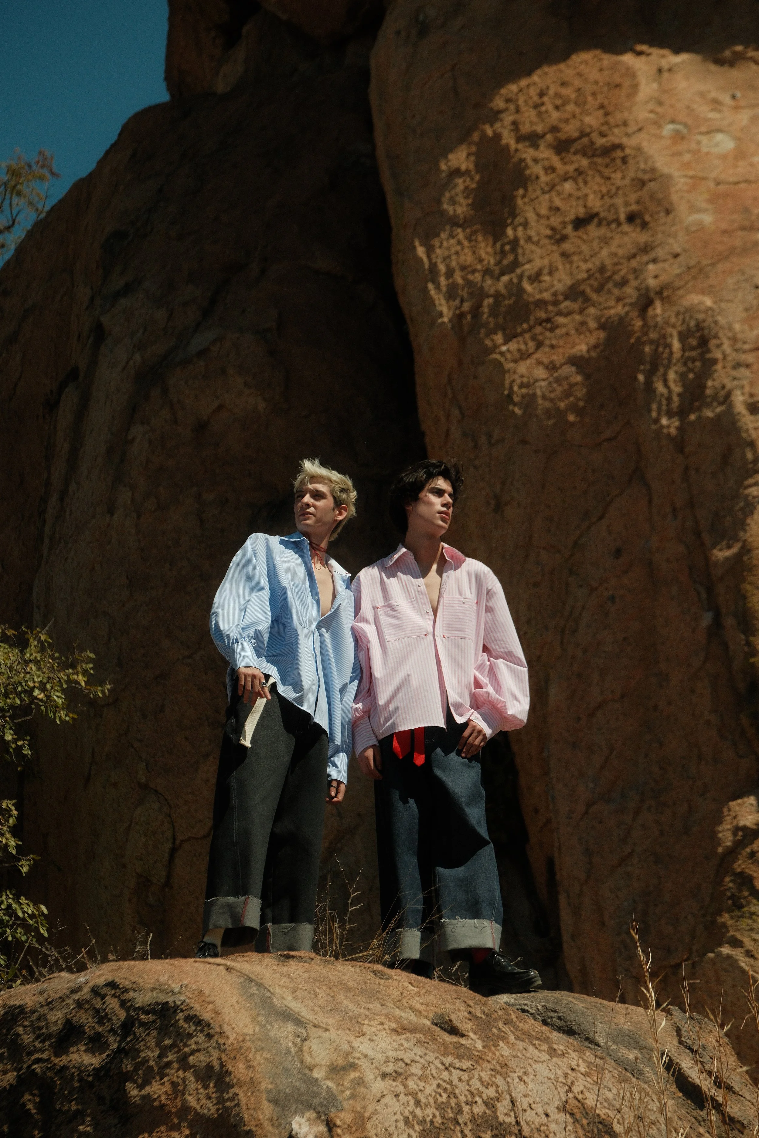 Dos personas con ropa informal y pantalones anchos, una con camiseta azul y otra rosa, de pie en una roca frente a una formación rocosa.