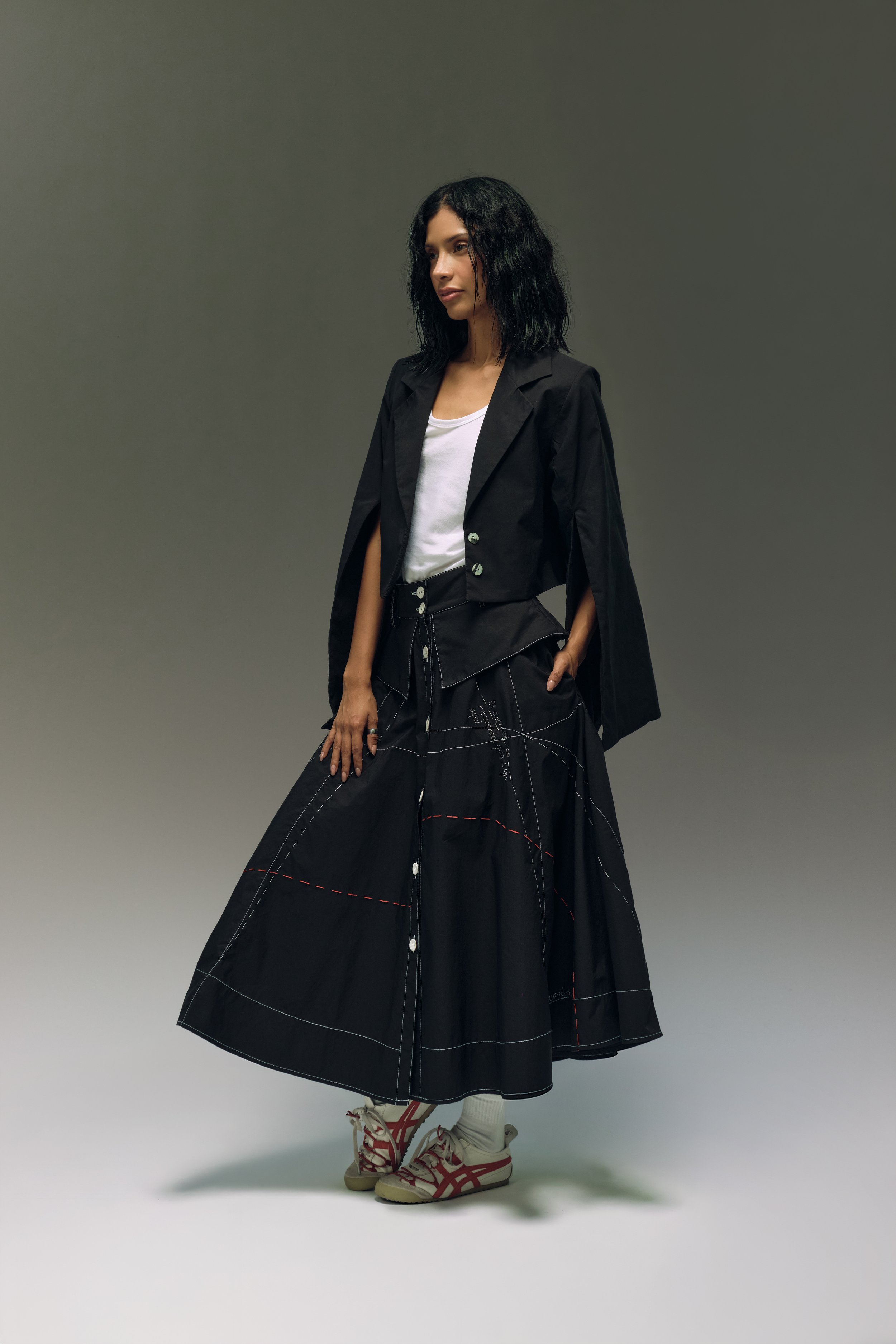 Mujer con cabello rizado y oscuro, vestida con chaqueta negra, falda larga a cuadros y zapatillas blancas con rojo, posando en un fondo gris.