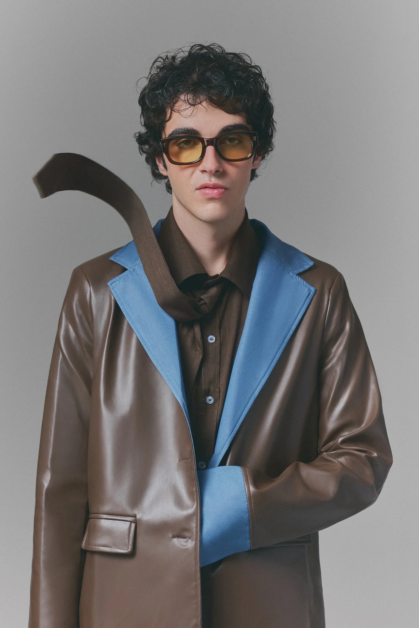 Retrato de un hombre joven con cabello rizado oscuro, gafas de sol de color ámbar, ropa elegante con chaqueta de cuero de color marrón y azul, y una camisa negra, sosteniendo una manguera o un tubo en el hombro izquierdo, contra un fondo gris simple.