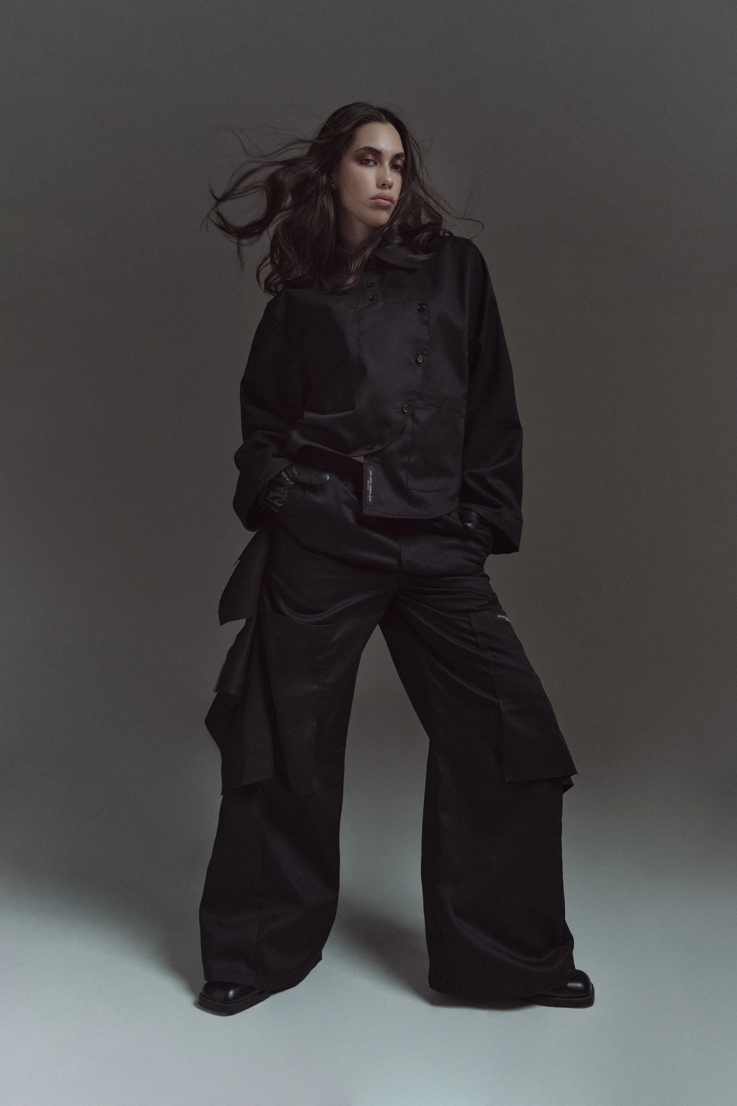 Mujer con cabello oscuro y rizado, vestida con ropa negra asimétrica, de pie en un fondo gris oscuro.