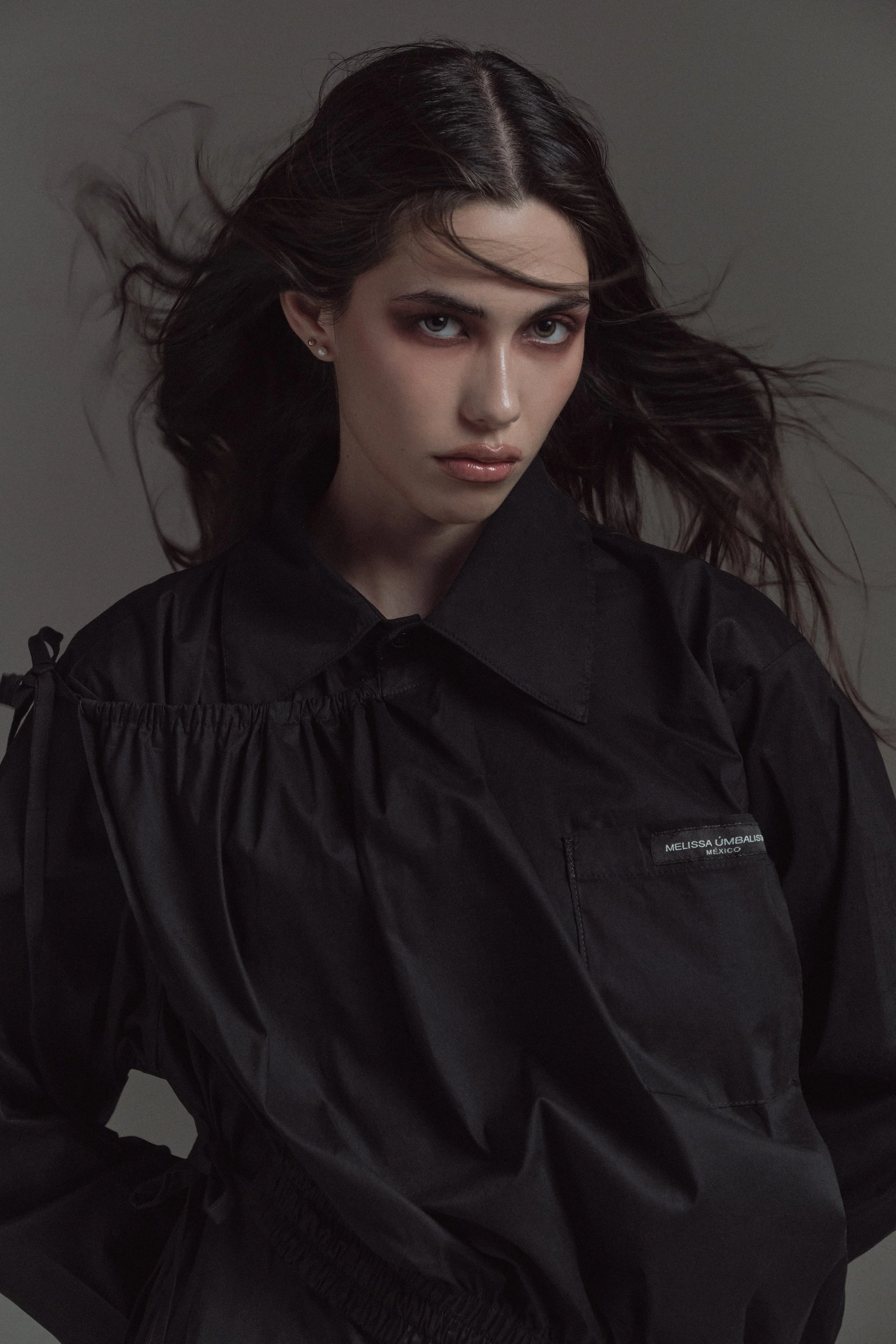 Retrato de una mujer joven con cabello oscuro, peinado hacia atrás, con expresión seria, usando una chaqueta negra con etiqueta que dice 'Melissa Umbal, México', en un fondo gris.