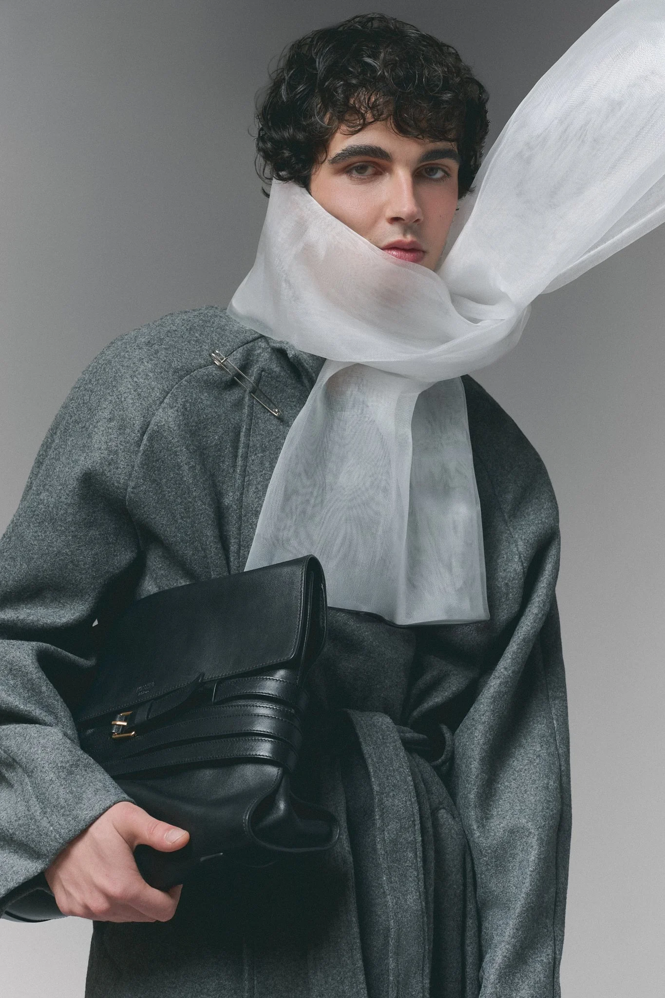 Joven con cabello rizado oscuro, vestido con un abrigo gris y bufanda blanca, sosteniendo una cartera negra y un bolso, con fondo gris uniforme.