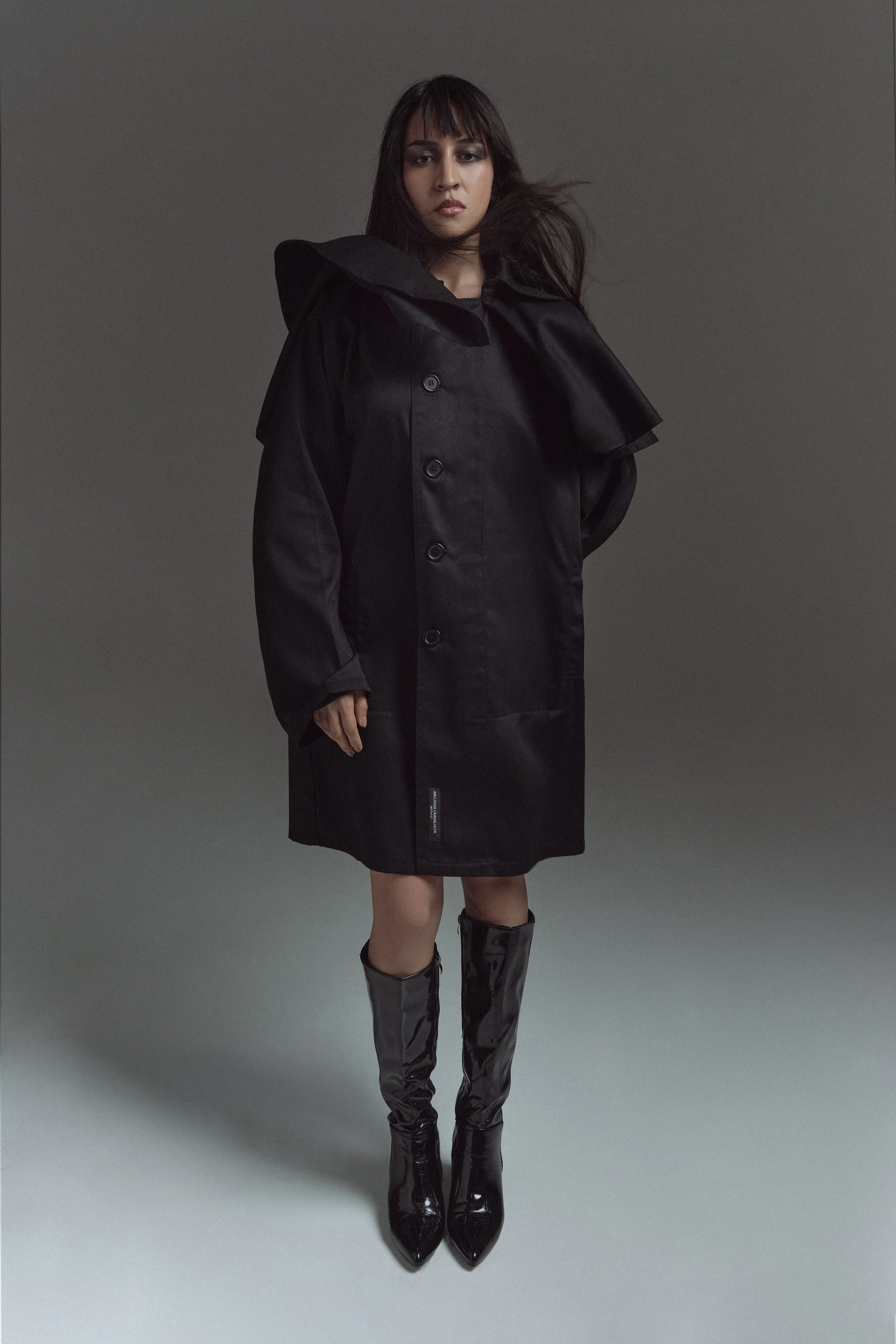 Mujer vestida con abrigo largo negro, botas altas negras de charol y cabello suelto, en fondo gris oscuro.