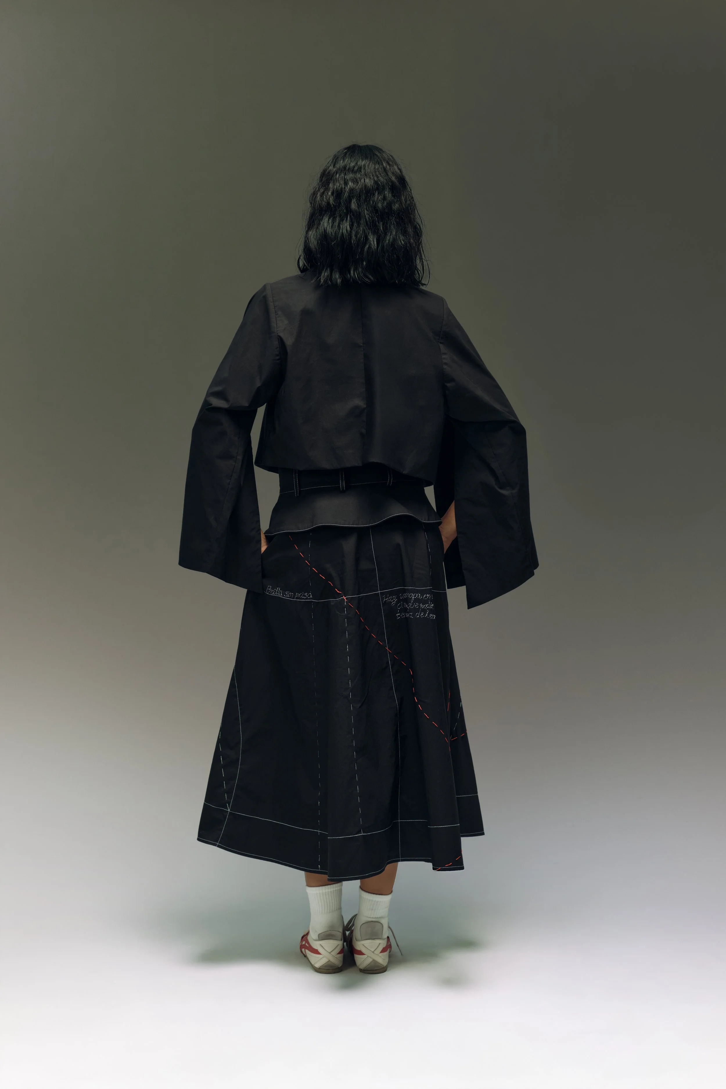 Persona de espaldas con pantalón largo negro, camiseta ajustada y chaqueta negra, calza deportivas y calcetas blancas, en un fondo gris sin detalles.