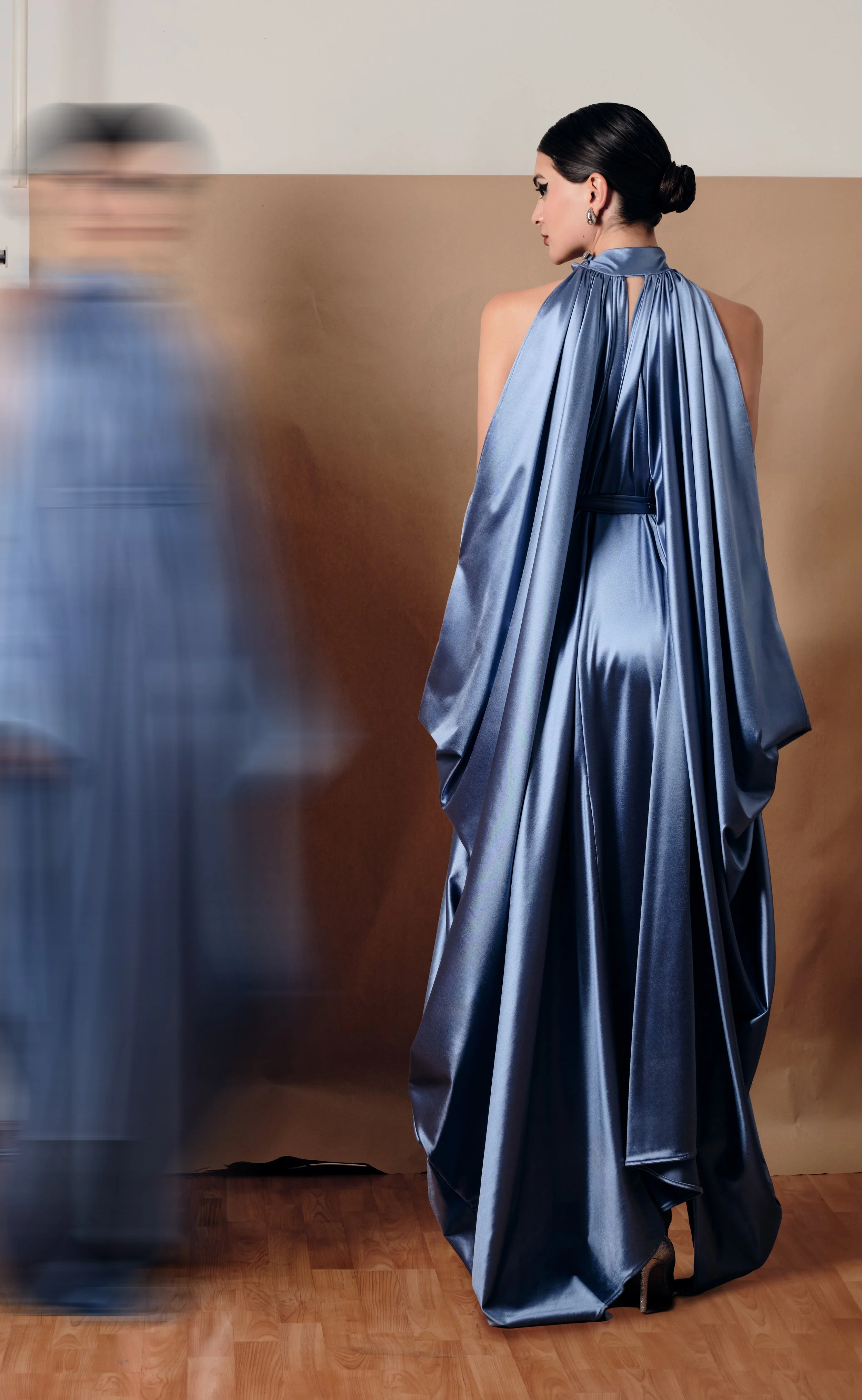 Una mujer con vestido largo y brillante en color azul, con el cabello peinado en un moño, de perfil, que parece estar en un evento o sesión de fotos, con un fondo simple y un suelo de madera.